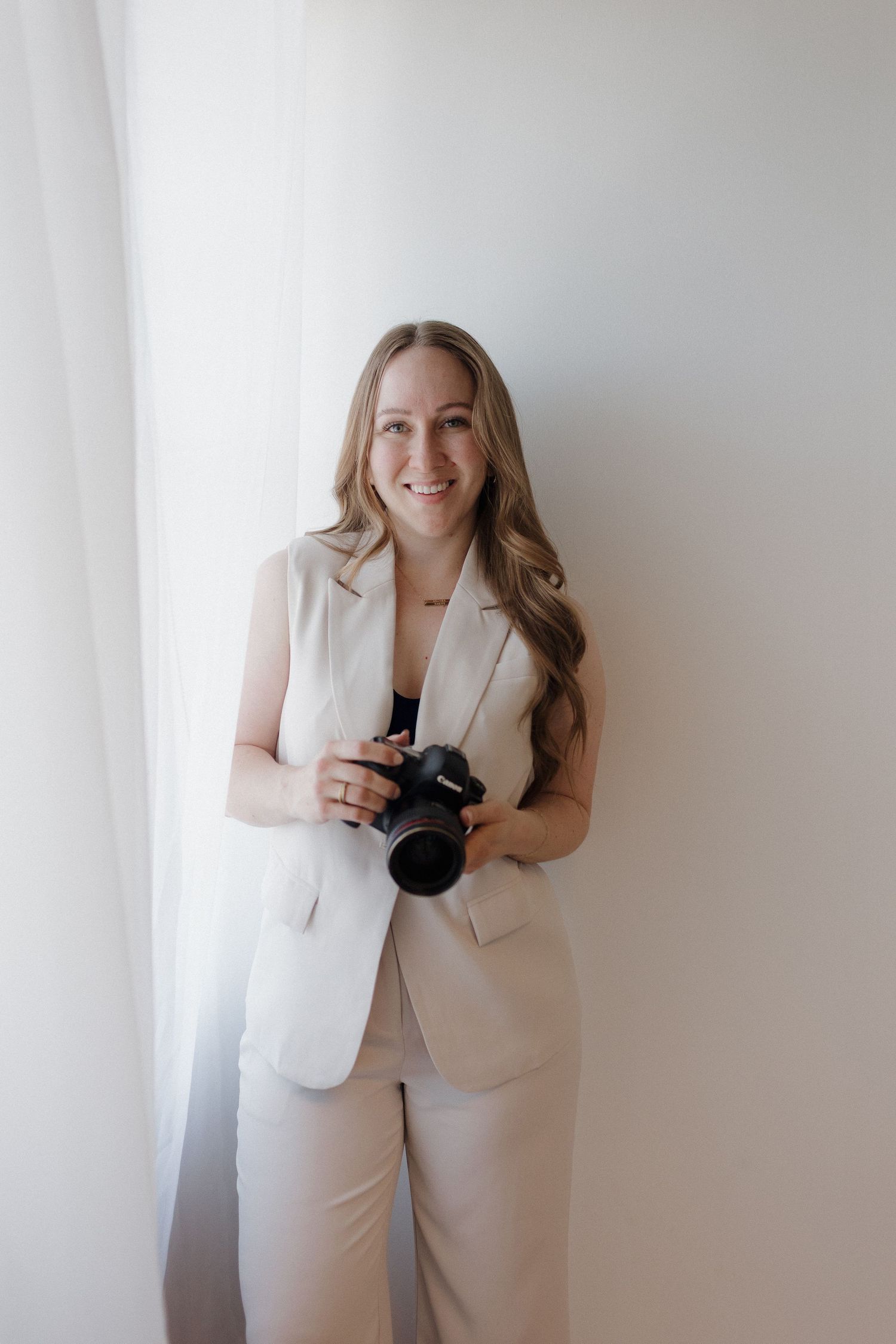 A photographer in an all-white pantsuit poses for professional headshots against a white backdrop.