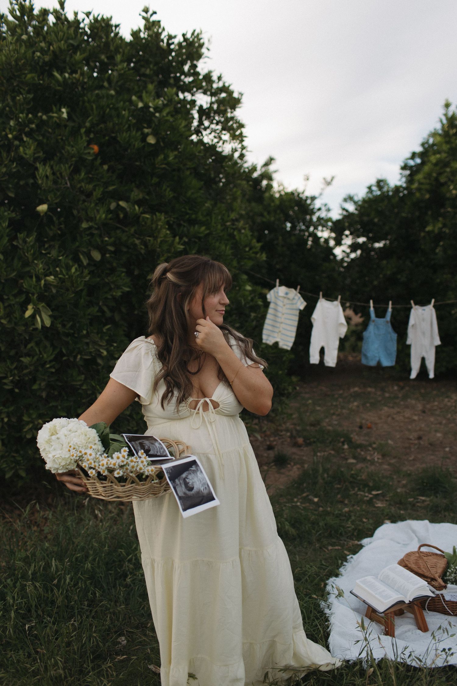 Pregnant woman in white dress holding basket with ultrasound photos and flowers, baby clothes hanging on line outdoors.