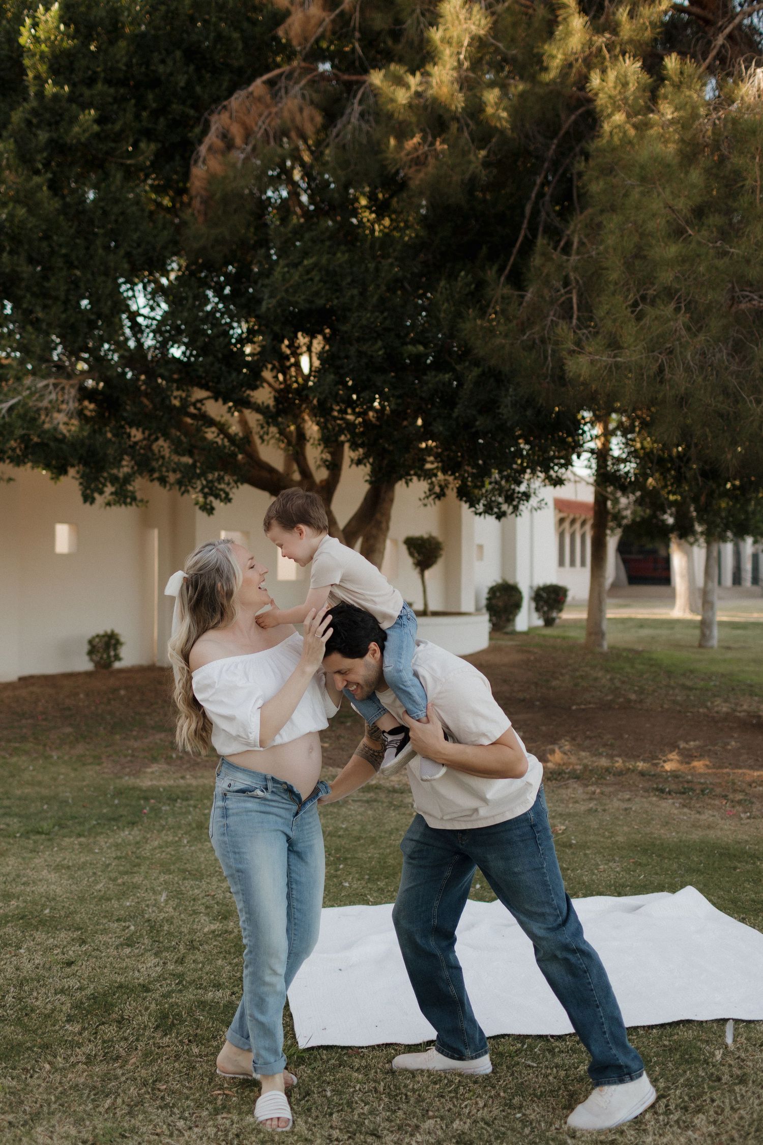 Playful family moment captured on white blanket under large tree at sunset.