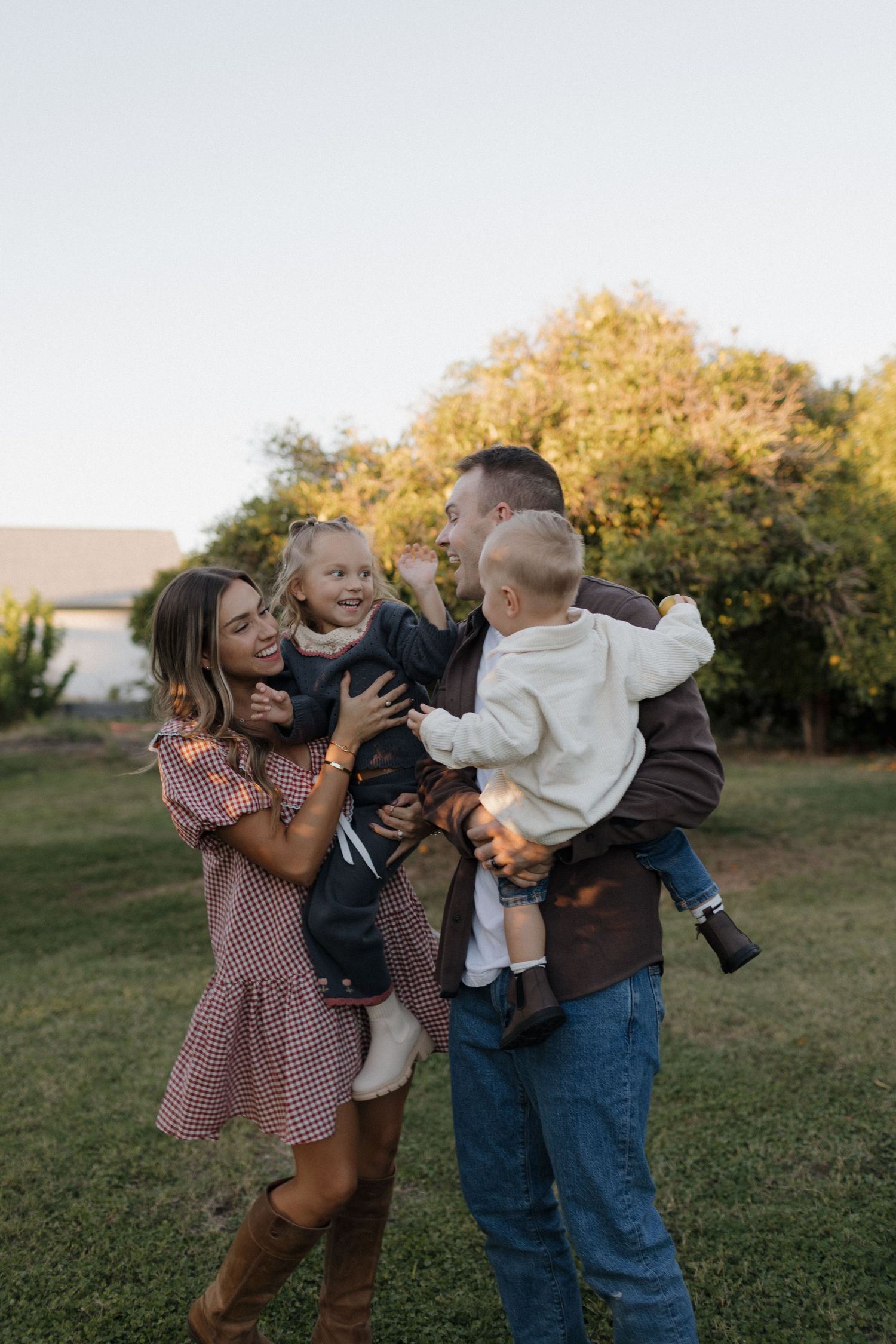 Happy family of four laughing outdoors, parents holding two young children in a sunny green backyard setting.