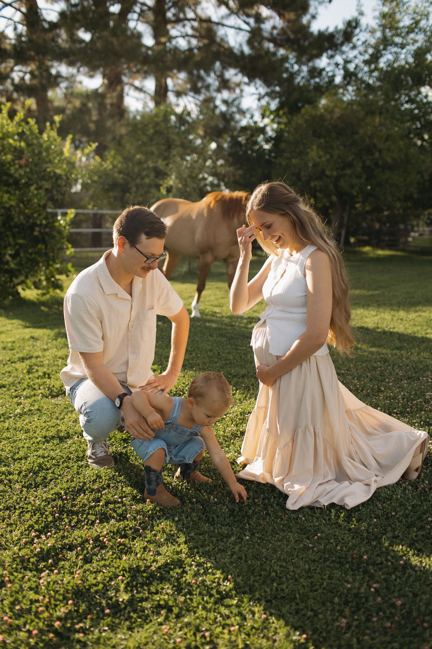A family with a toddler enjoys a sunny day on green grass with a brown horse grazing in the background.