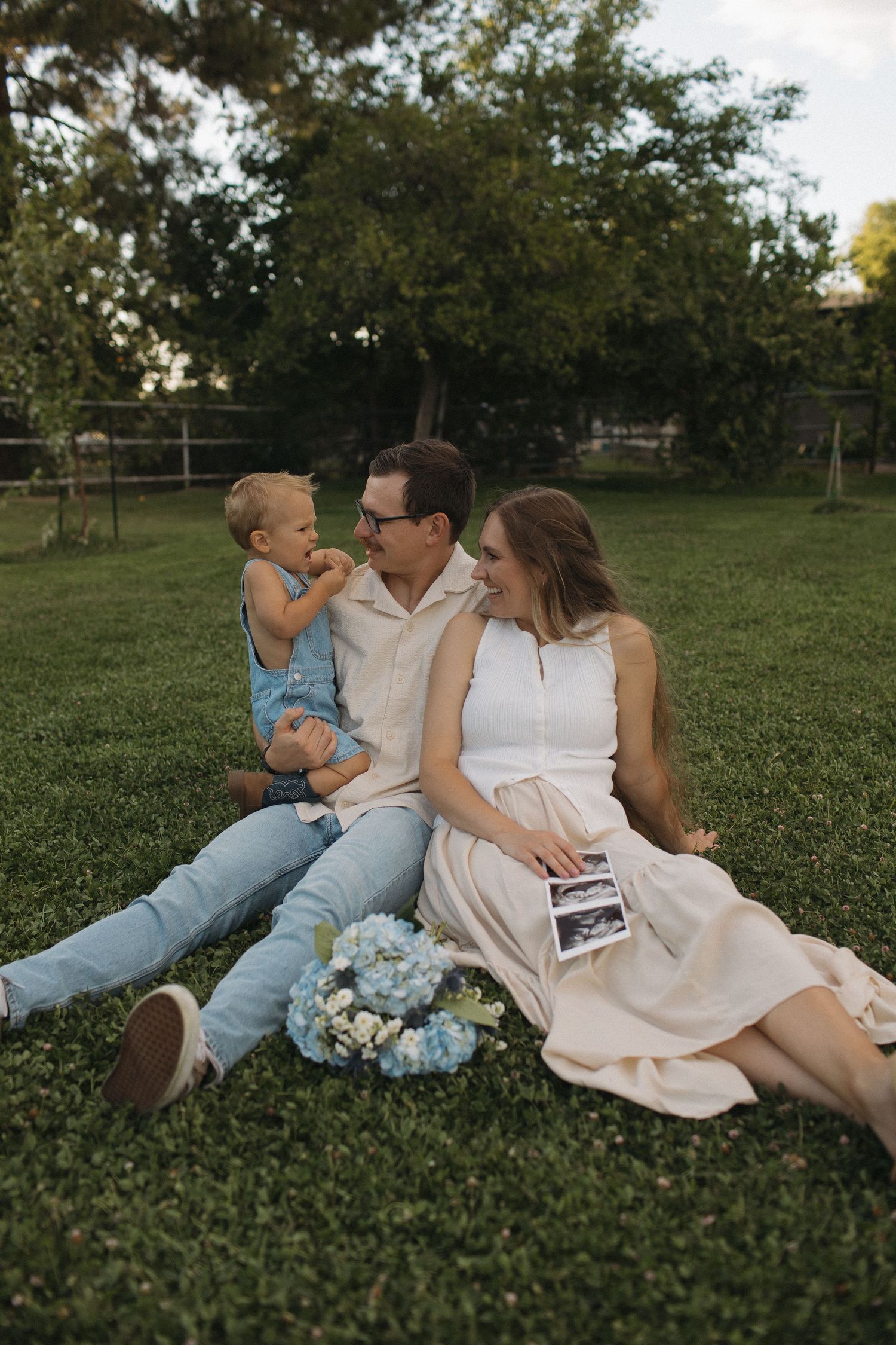 Pregnant woman in white dress sits on grass with husband and toddler, holding ultrasound photo, blue hydrangeas nearby.
