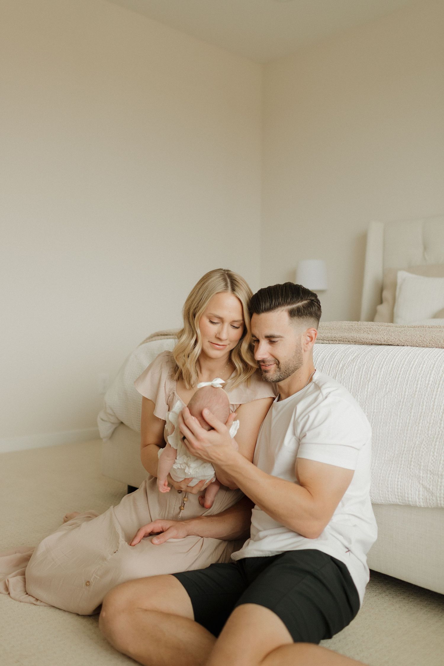A couple sits together on a light colored floor holding their newborn baby girl in an intimate indoor setting.