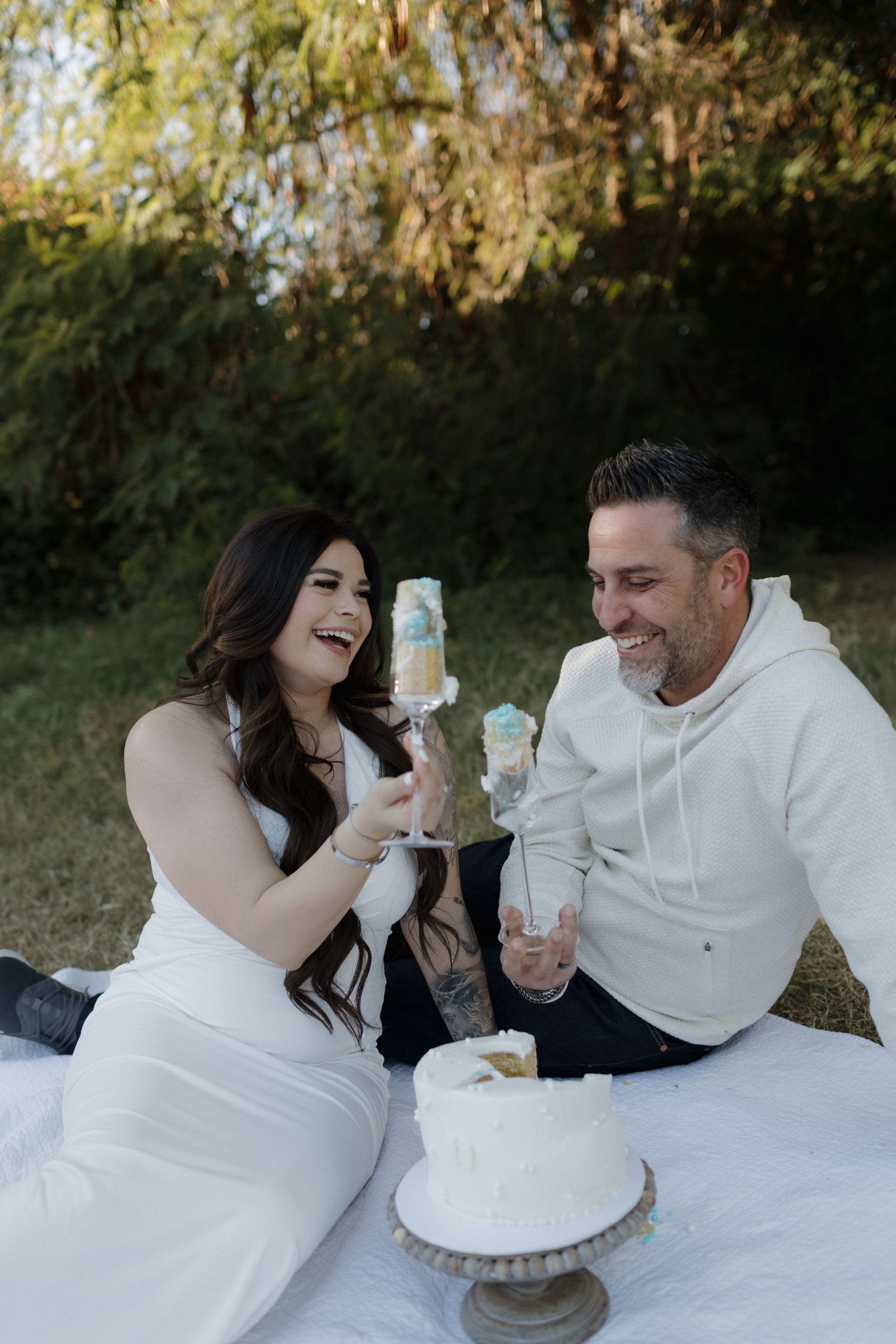 Playful couple in white attire share cake and laughter during an outdoor photo session on a white blanket.