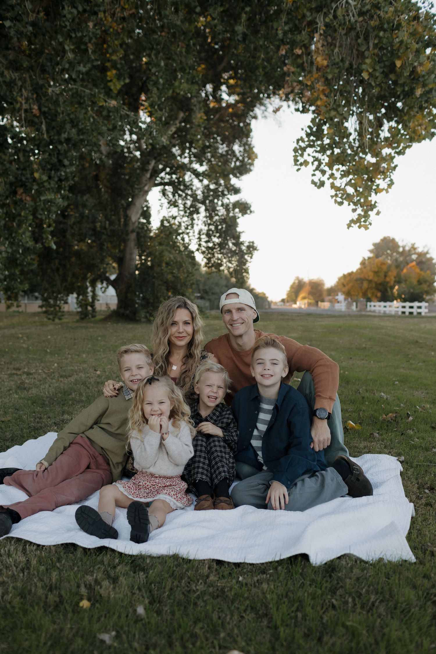 Happy family of six poses on a white blanket under a large tree during an outdoor fall photo session.