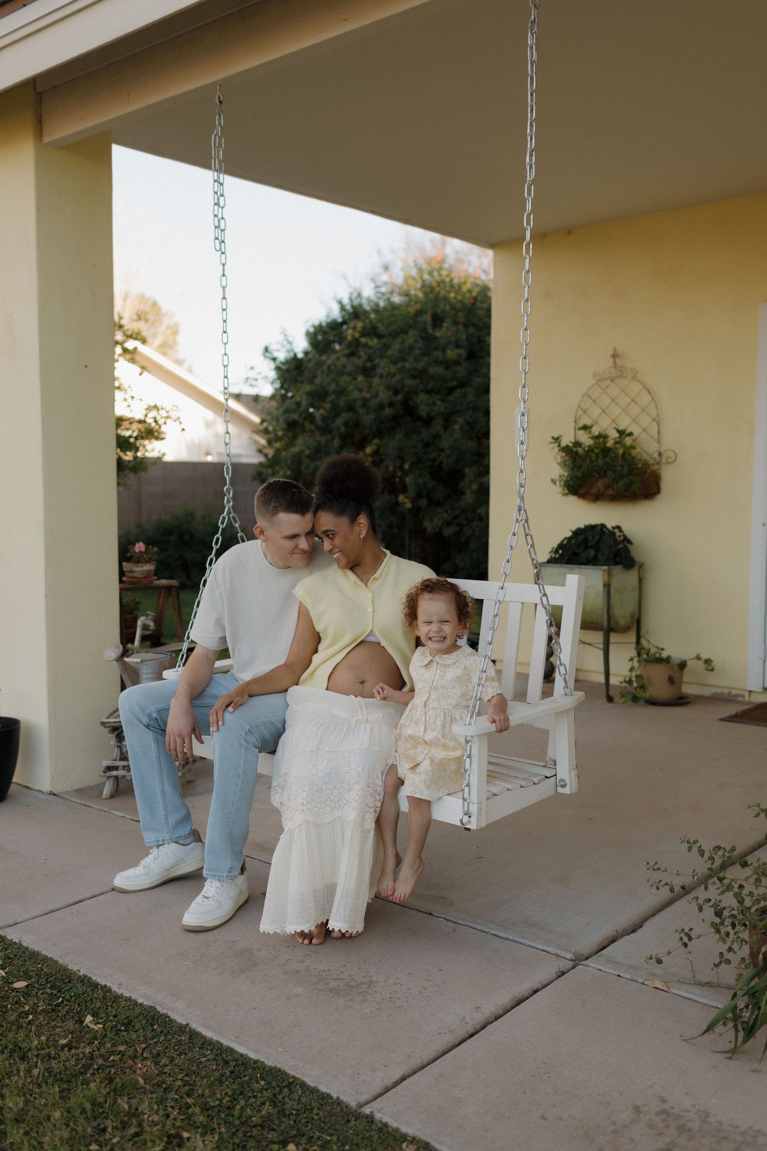 A family of three sits on a white porch swing, with a pregnant mother, father, and toddler enjoying a warm evening outside.