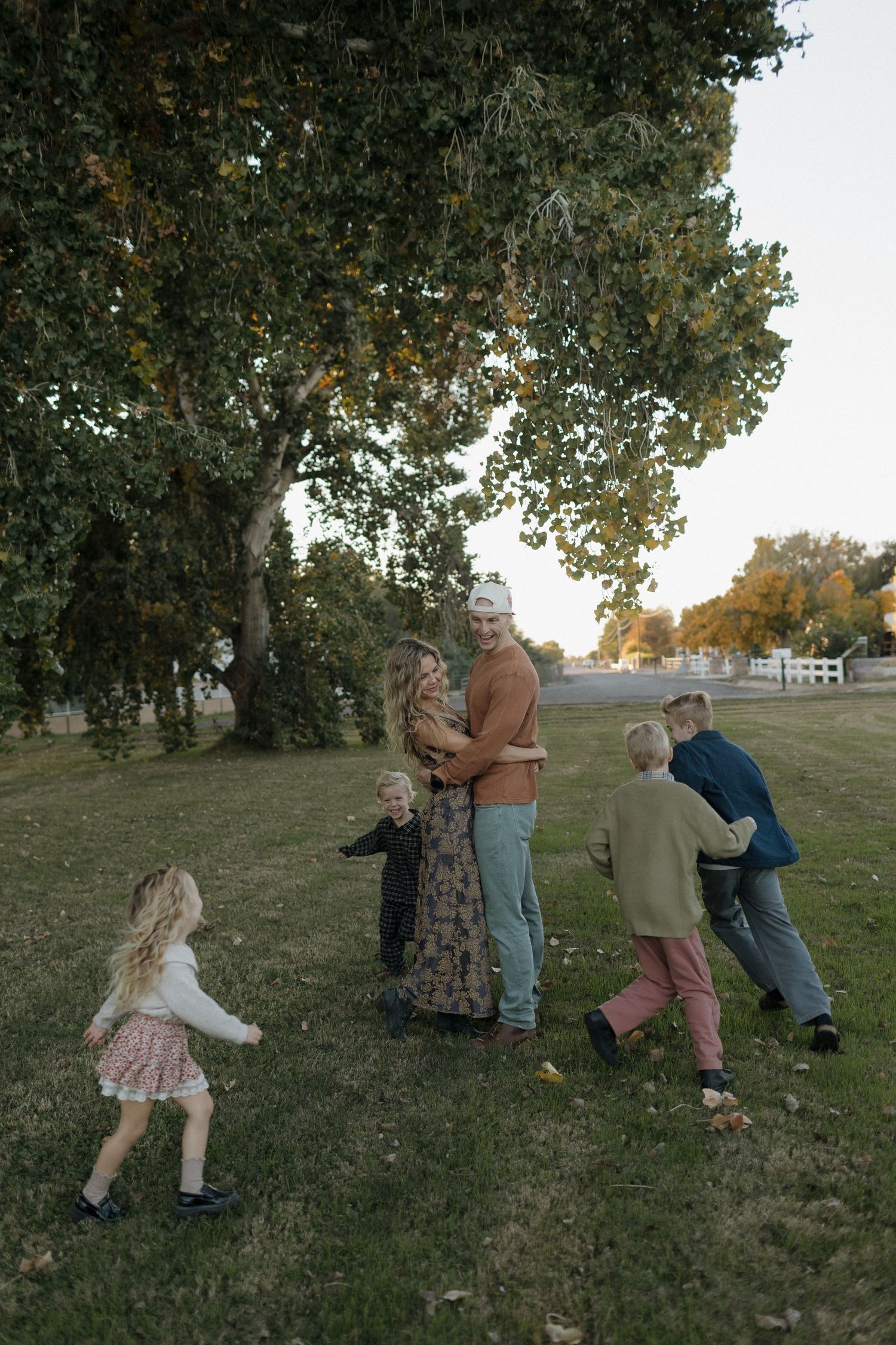 A group of people play a game of tag around a large tree in a grassy field on an autumn day.