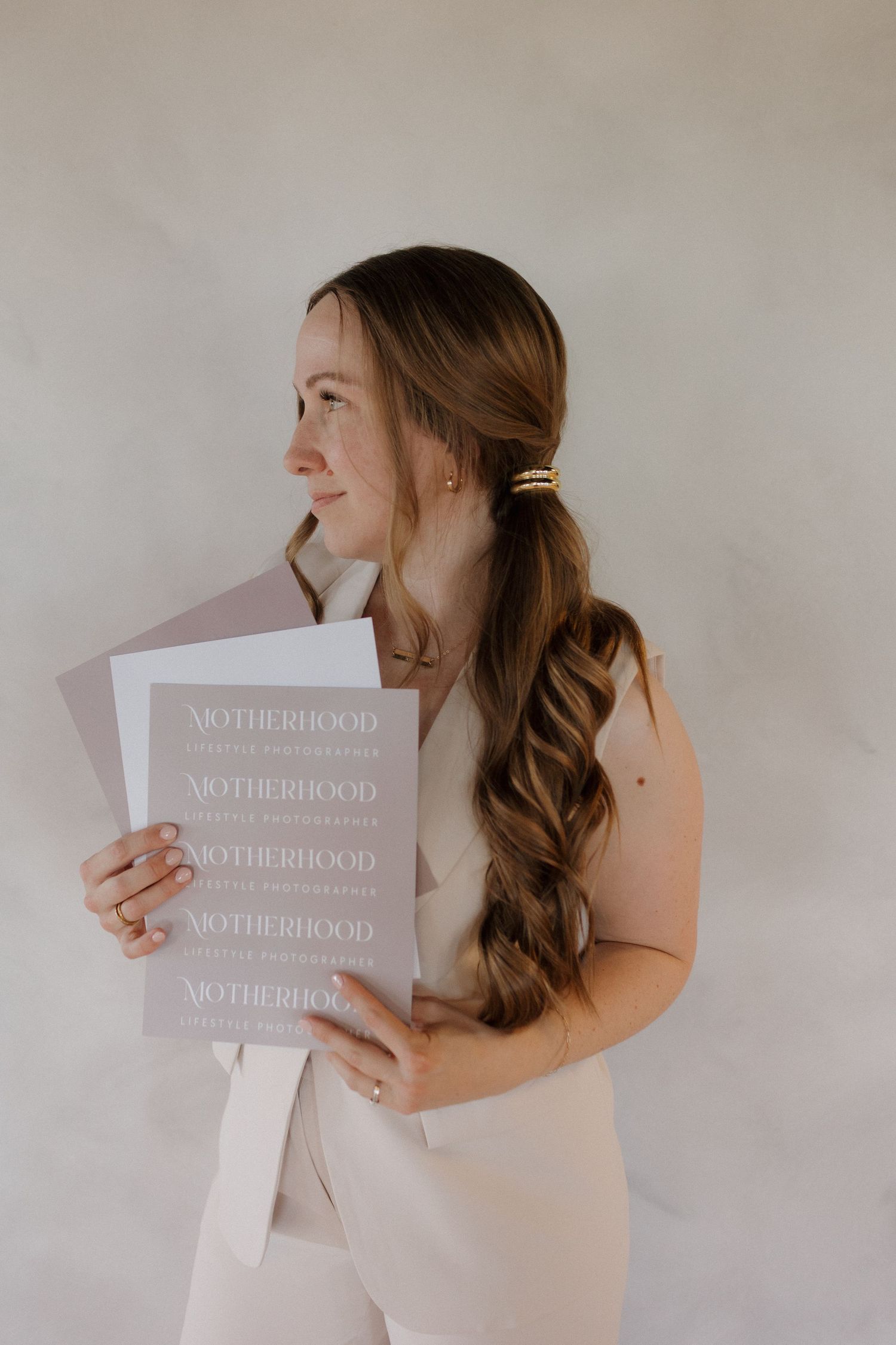 Photographer with long wavy brown hair in light beige outfit holding copies of branding documents titled Motherhood against white wall.