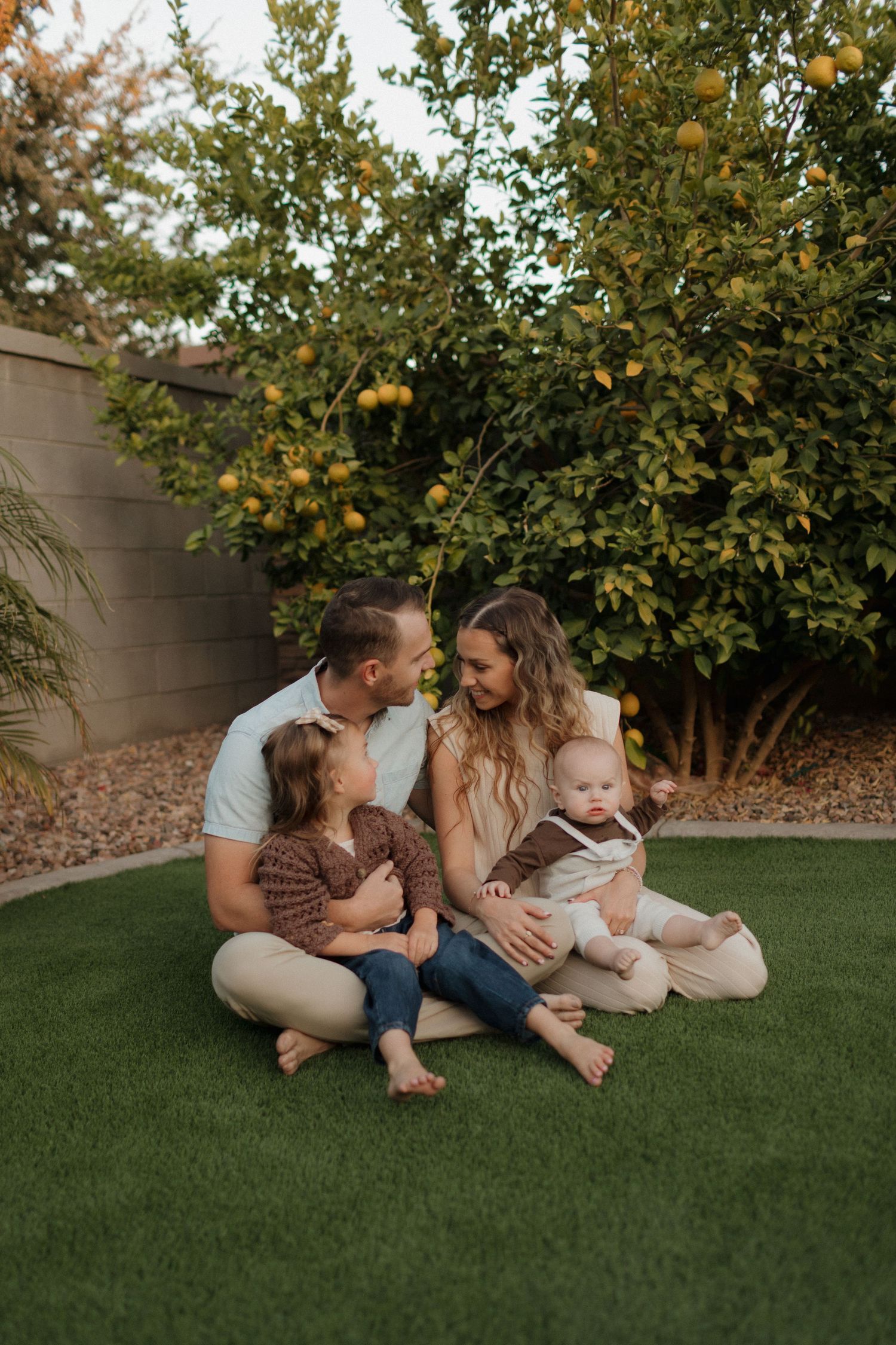 Family sits together on artificial grass beneath a citrus tree in their backyard.