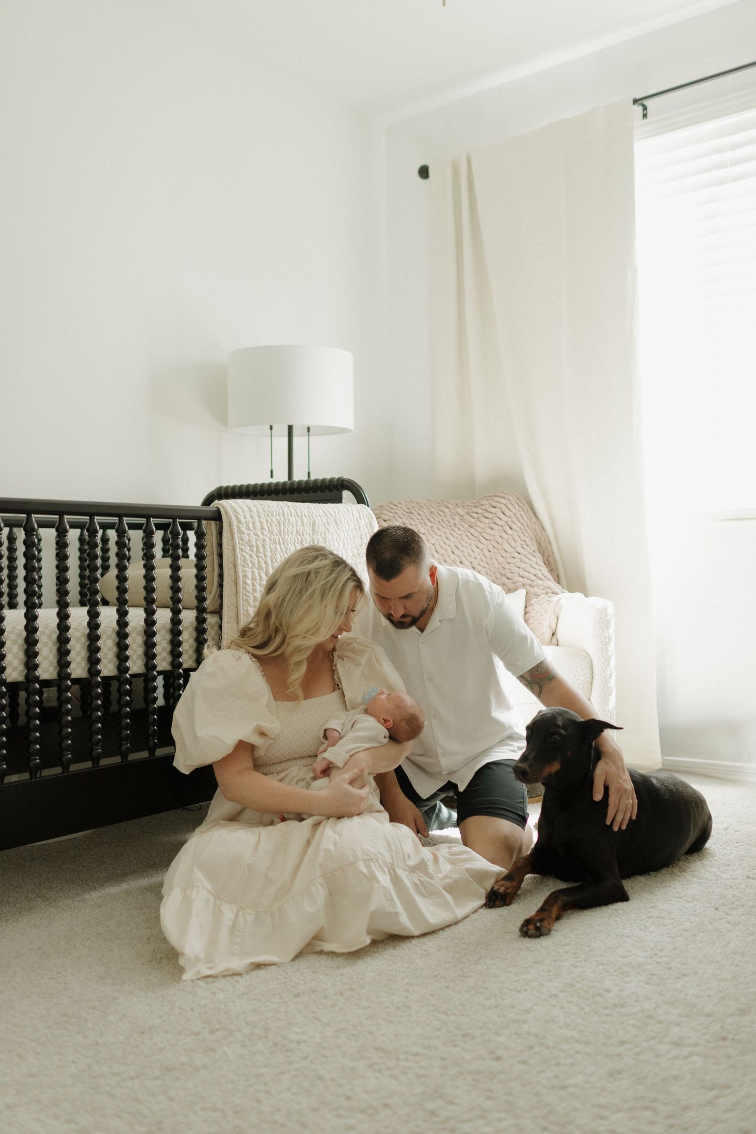 A cozy minimalist nursery scene with a black crib and family sitting on carpet with their dog.