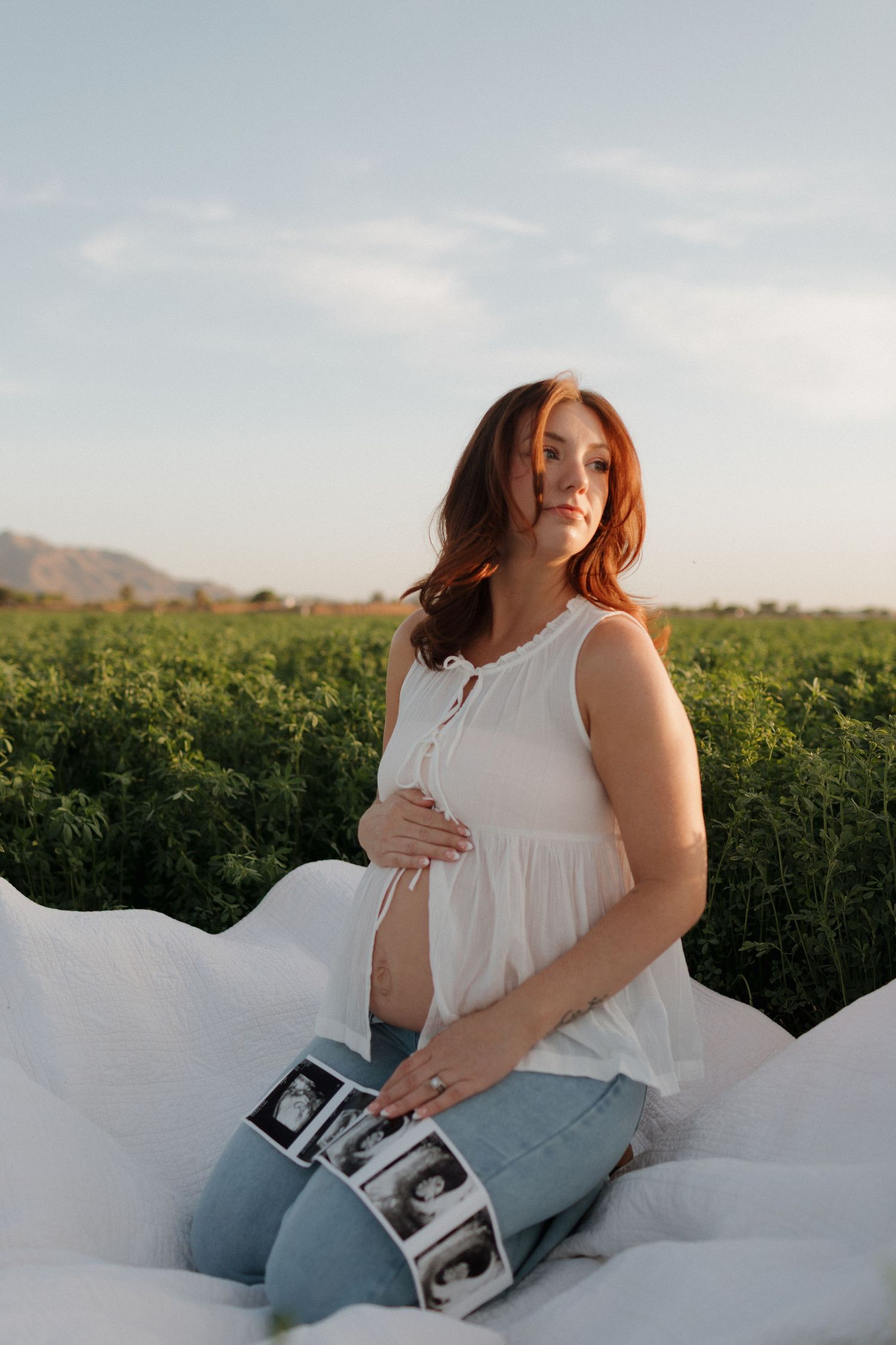 A maternity photo shoot in a green field at sunset with ultrasound photos and white fabric creating a dreamy scene.