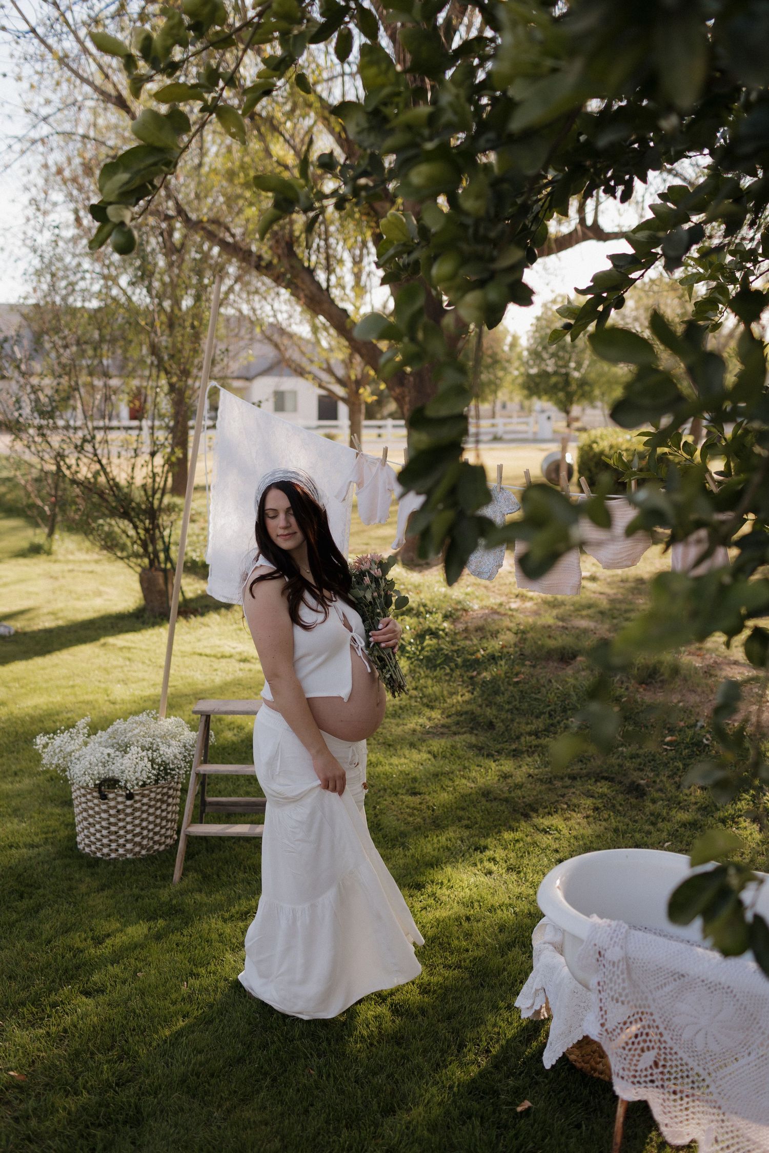 A person in a white dress stands in a sunlit garden with trees and white flowers in the background.