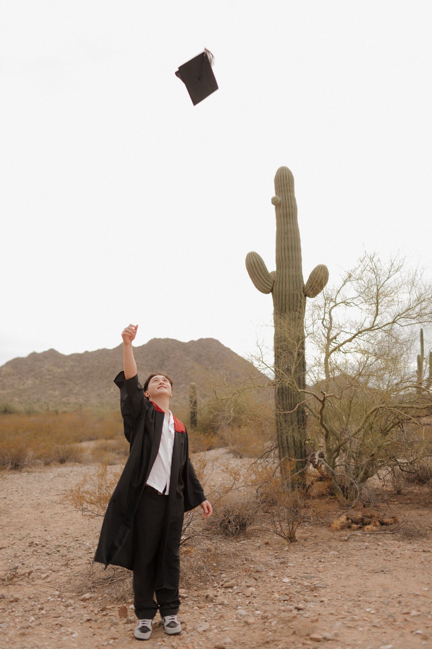 Graduate tossing cap in celebration near tall desert cactus.