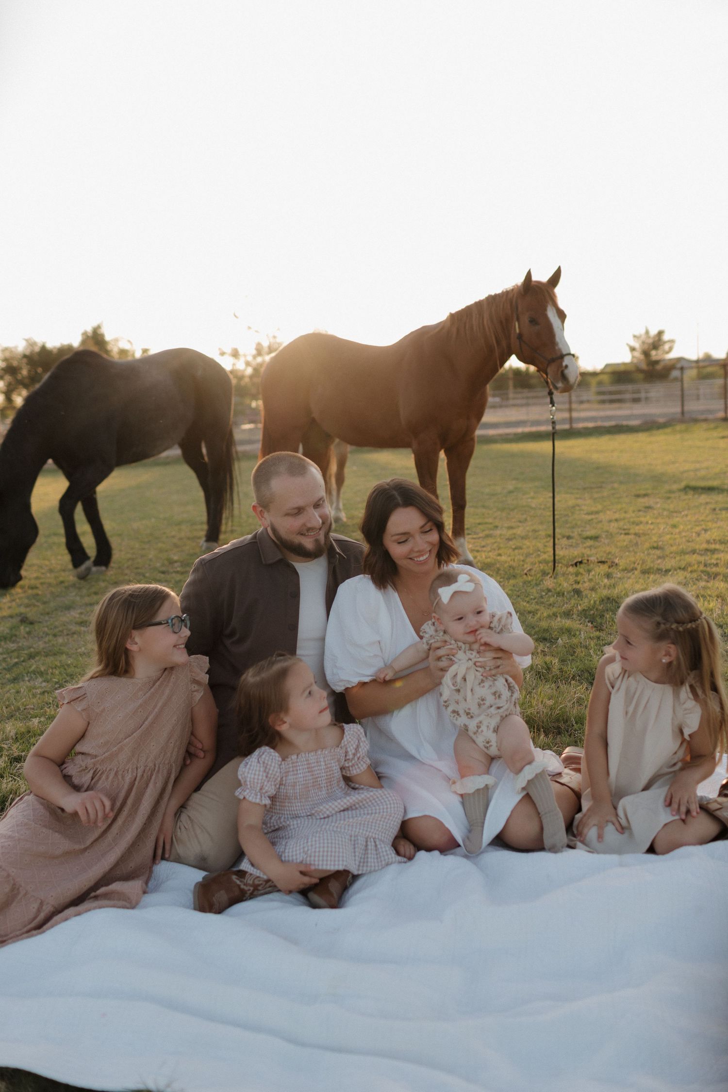 A joyful family relaxes together on a white blanket in a field with horses visible in the background at sunset.