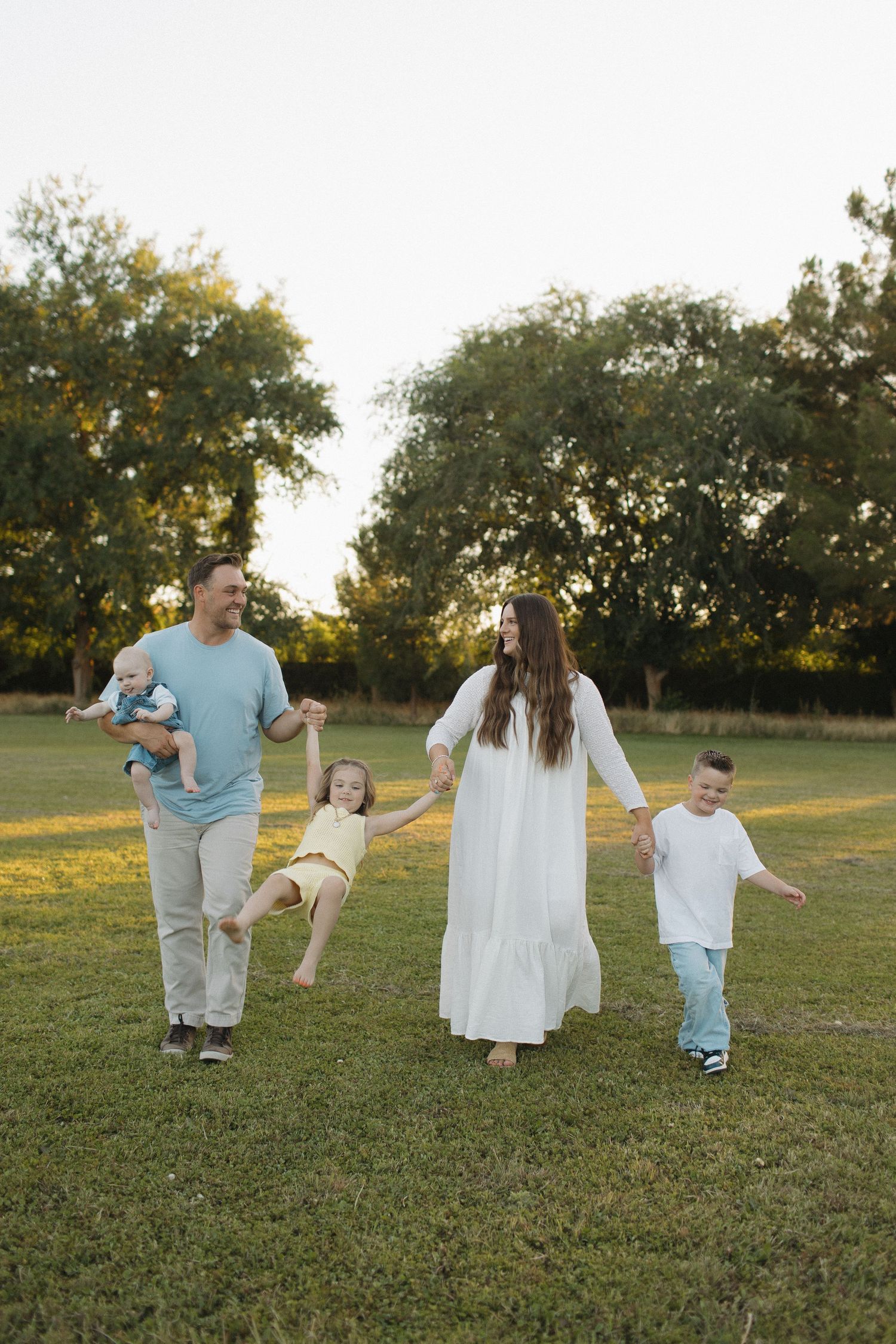 Happy family of five walking in a sunny green park, parents swinging daughter between them while holding hands outdoors.