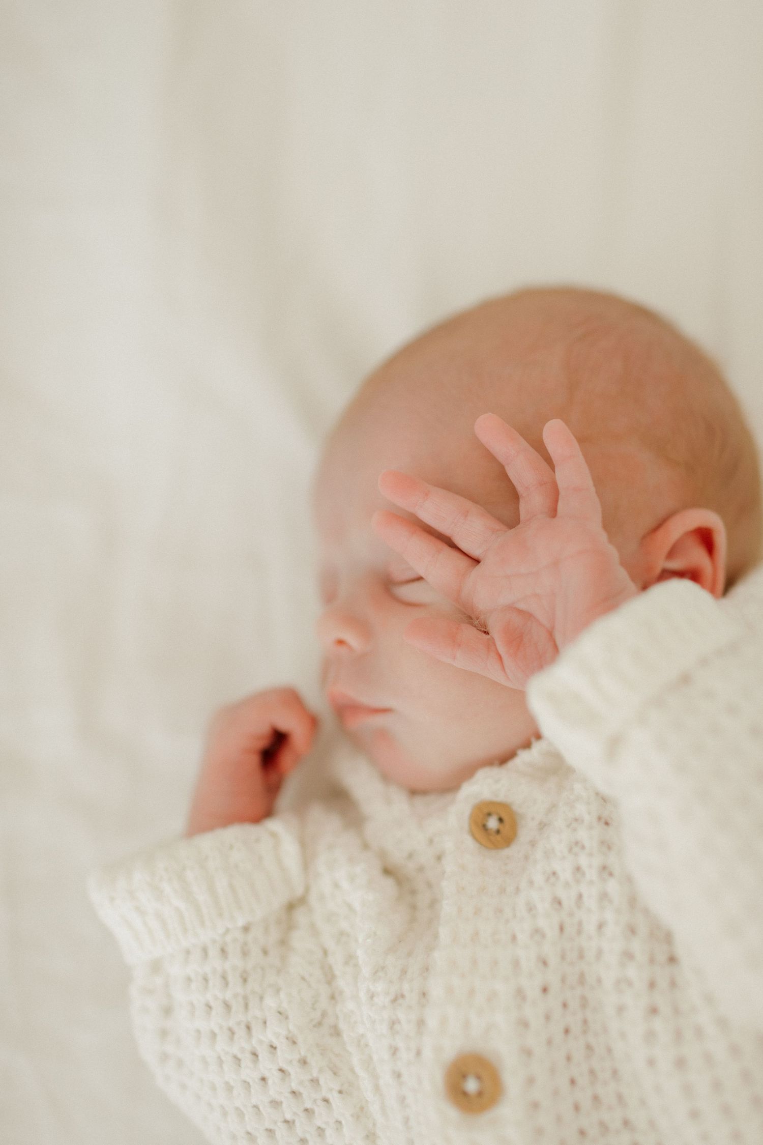 Close-up of a sleeping newborn in a white knit sweater with their tiny hand near their face.