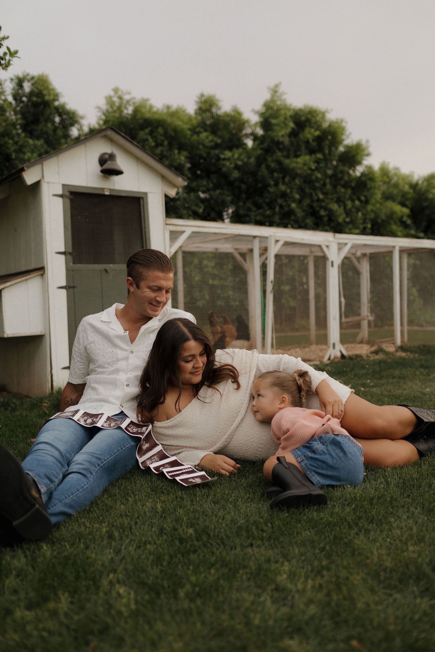 A happy family relaxes on green grass near a chicken coop, parents smiling at their young daughter.