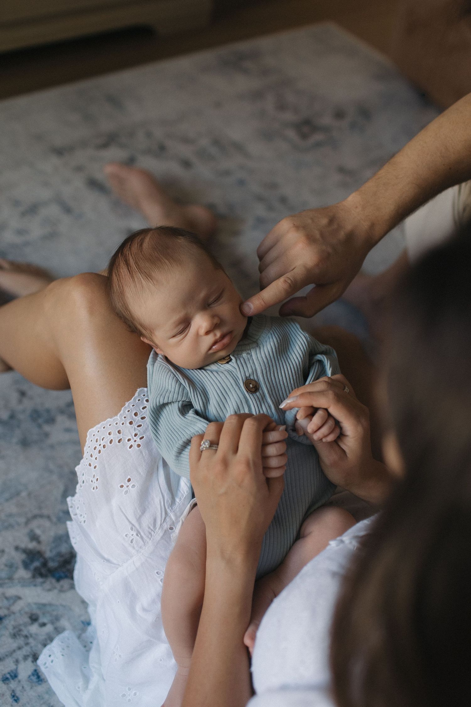 Parents tenderly holding their sleeping newborn baby dressed in a blue onesie on a soft blanket.