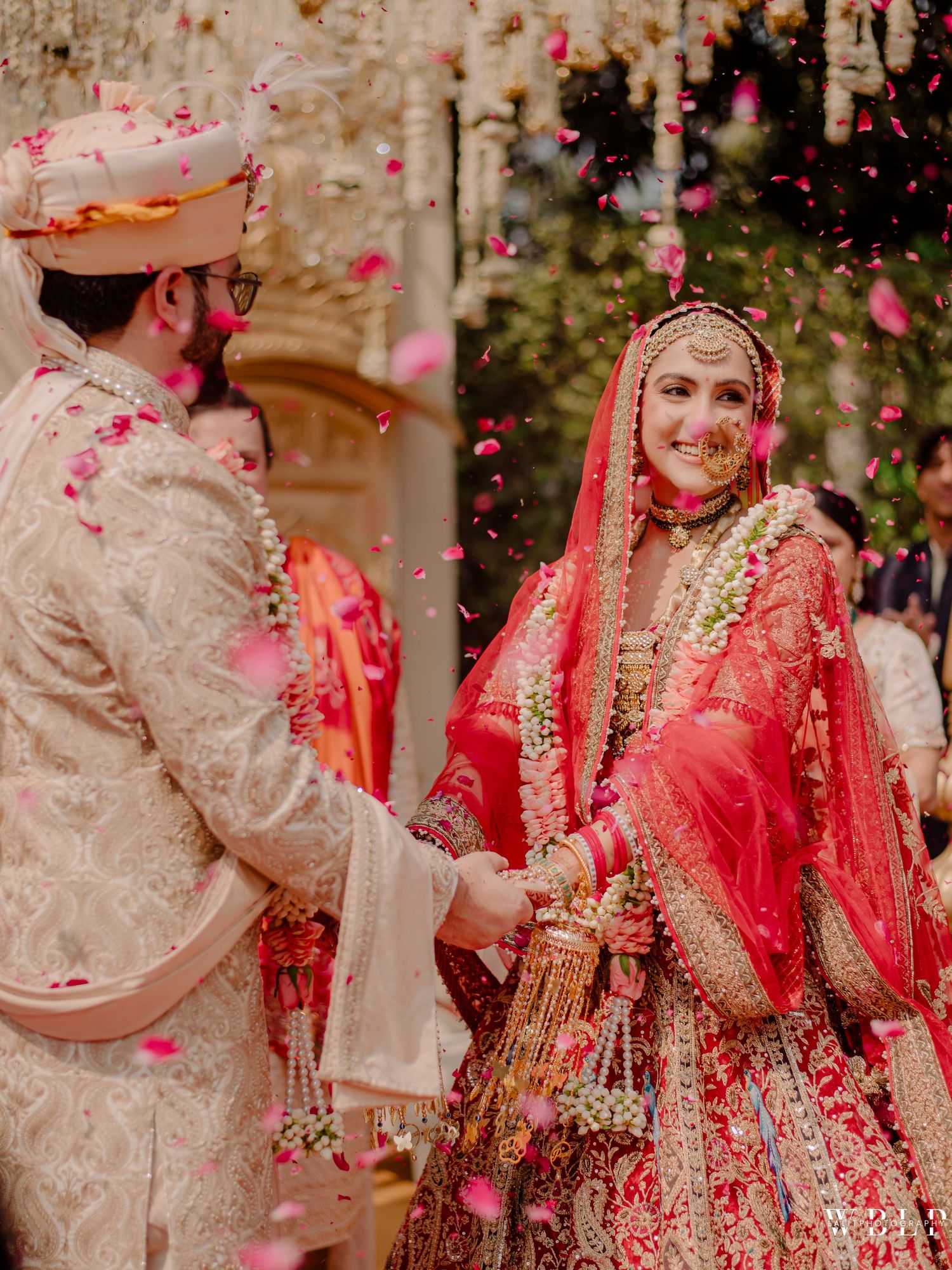 Indian bride and groom exchange garlands in traditional red lehenga and cream sherwani amid falling rose petals.