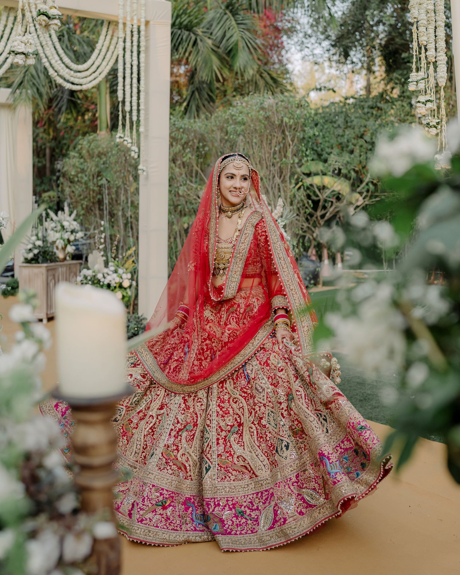 Bride in red and gold bridal lehenga with dupatta twirling at an outdoor wedding venue adorned with floral decorations.