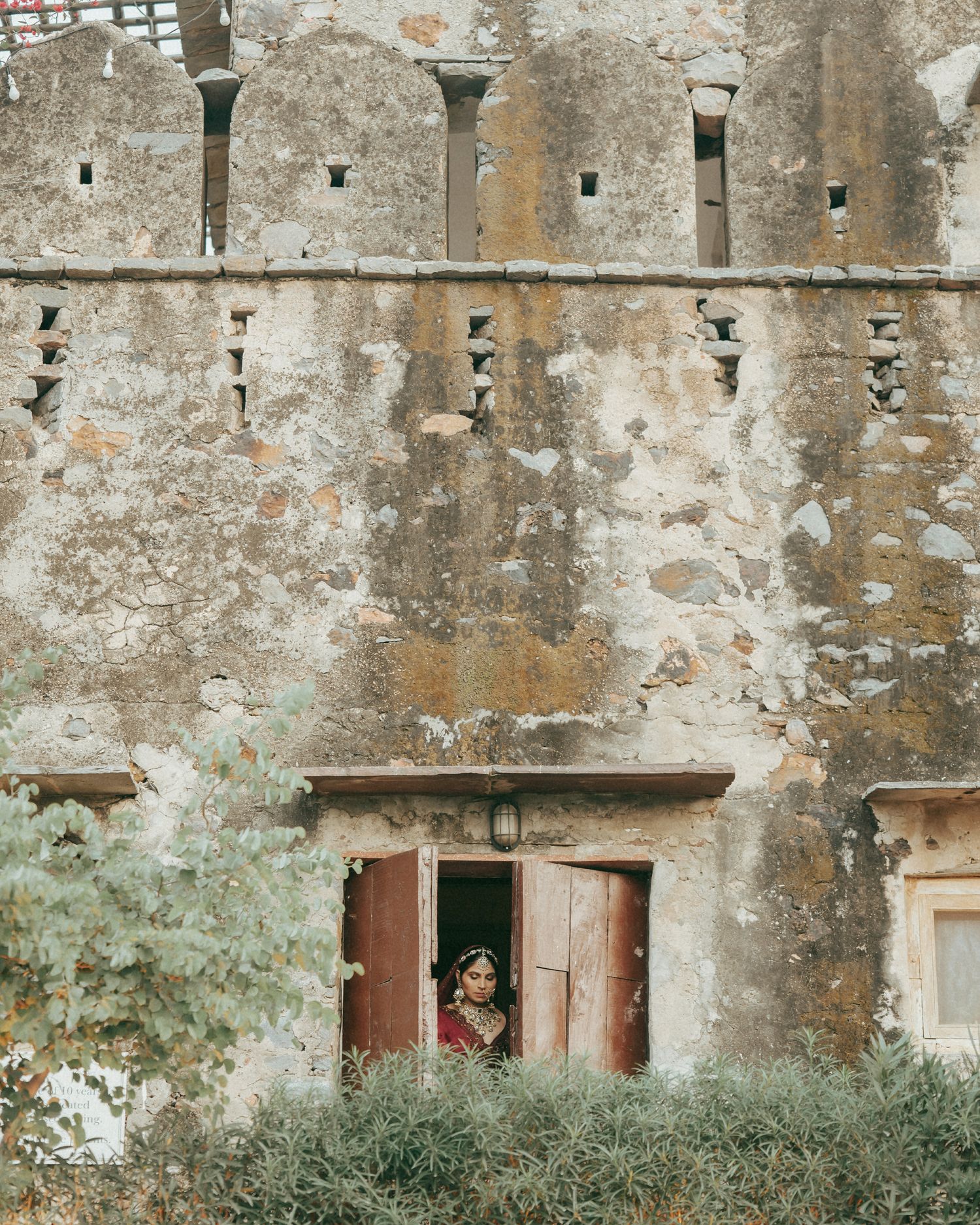 A bride in traditional Indian attire stands in a doorway of an ancient stone fort building surrounded by lush greenery.