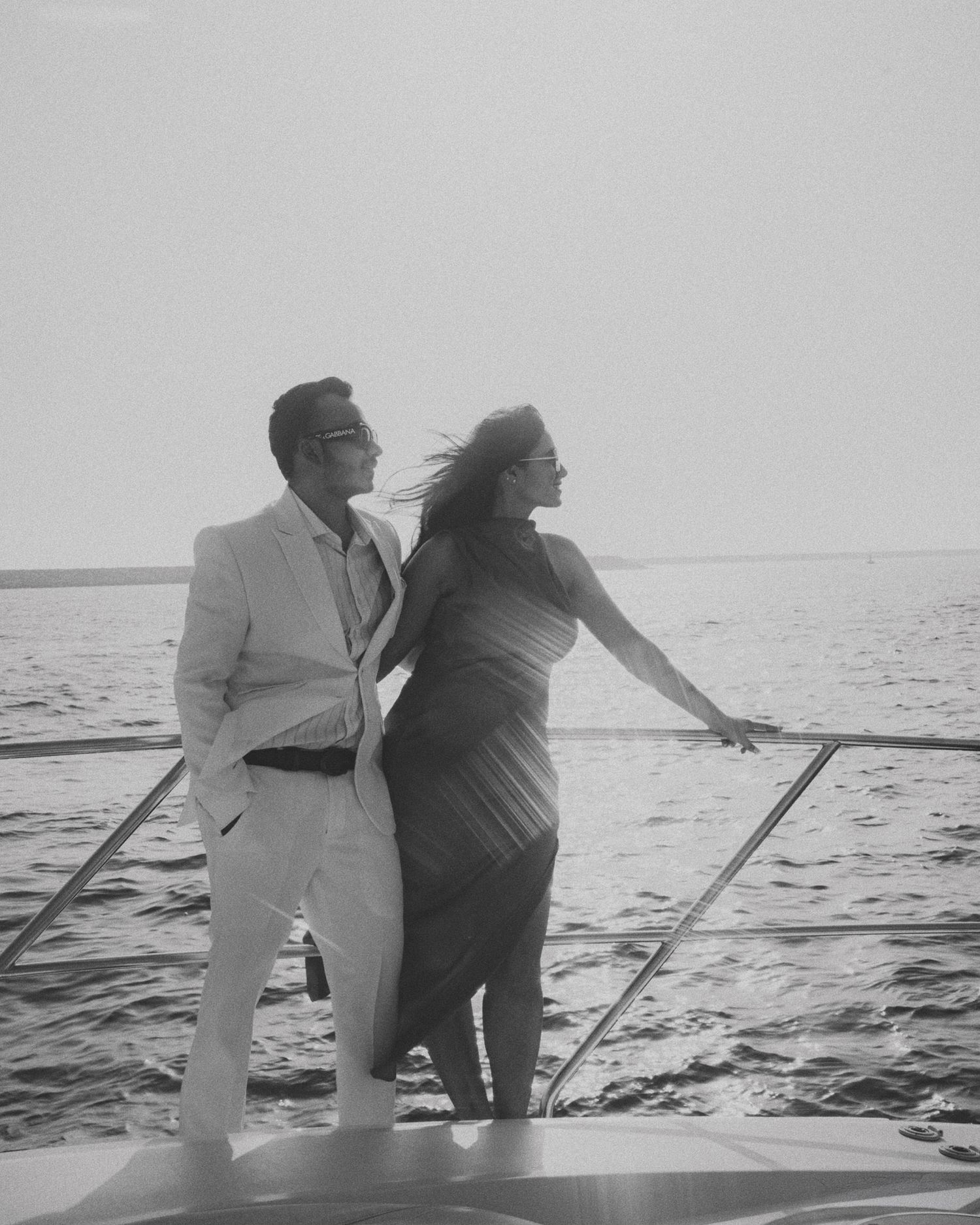 Elegant couple posing on luxury yacht bow in black and white, woman in draped dress, man in white suit, ocean backdrop.