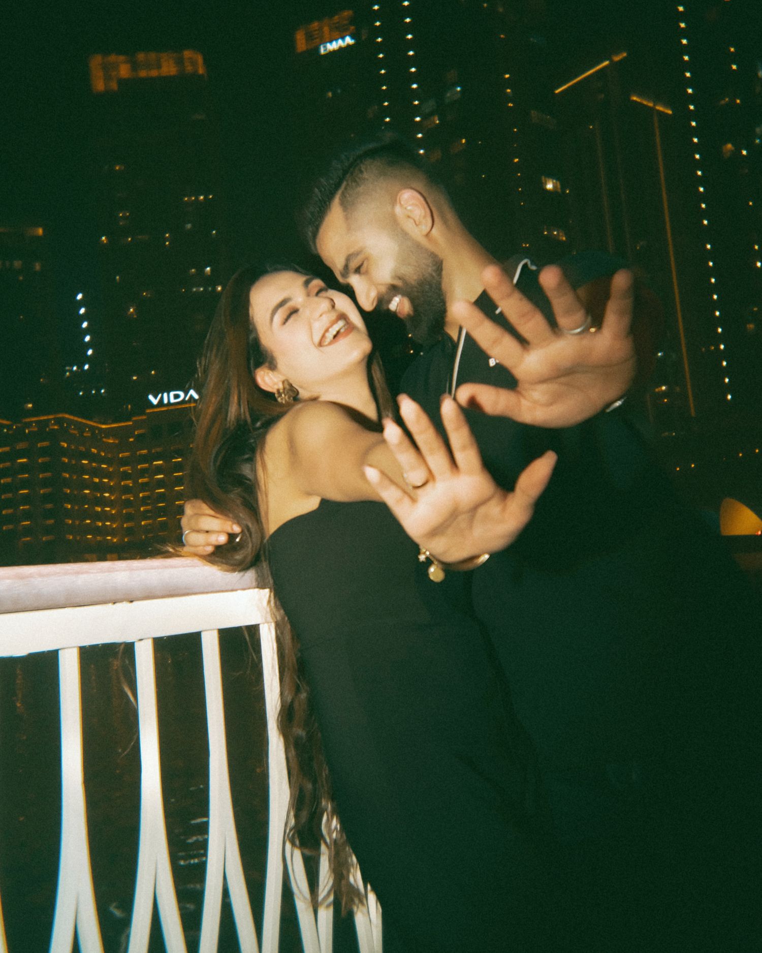 A couple in black outfits embraces and smiles on a rooftop at night with city lights glowing behind them.