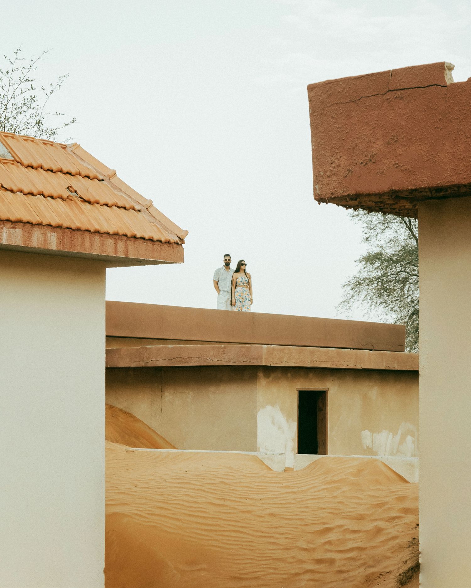 Couple standing on rooftop surrounded by sand dunes and terracotta buildings in a desert landscape.