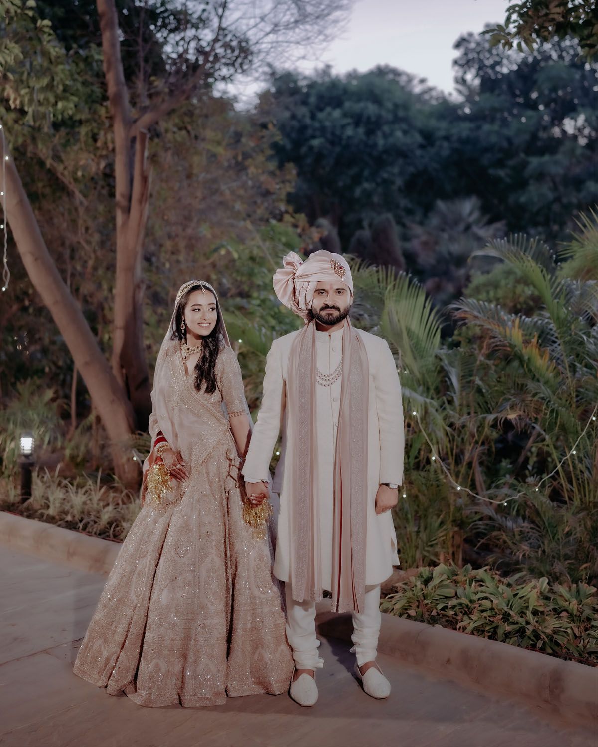 A couple in traditional South Asian wedding attire standing together in an outdoor garden setting.