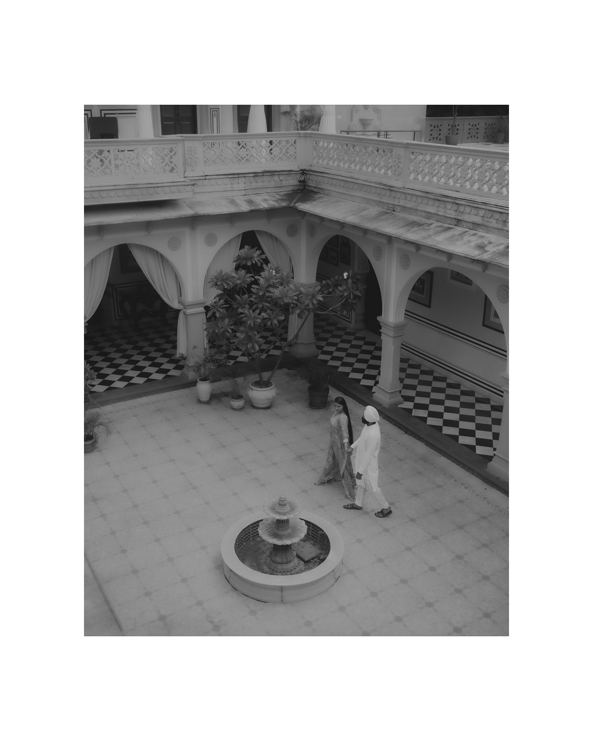 A historic courtyard with checkered flooring and arched colonnade features a central fountain and two figures standing nearby.