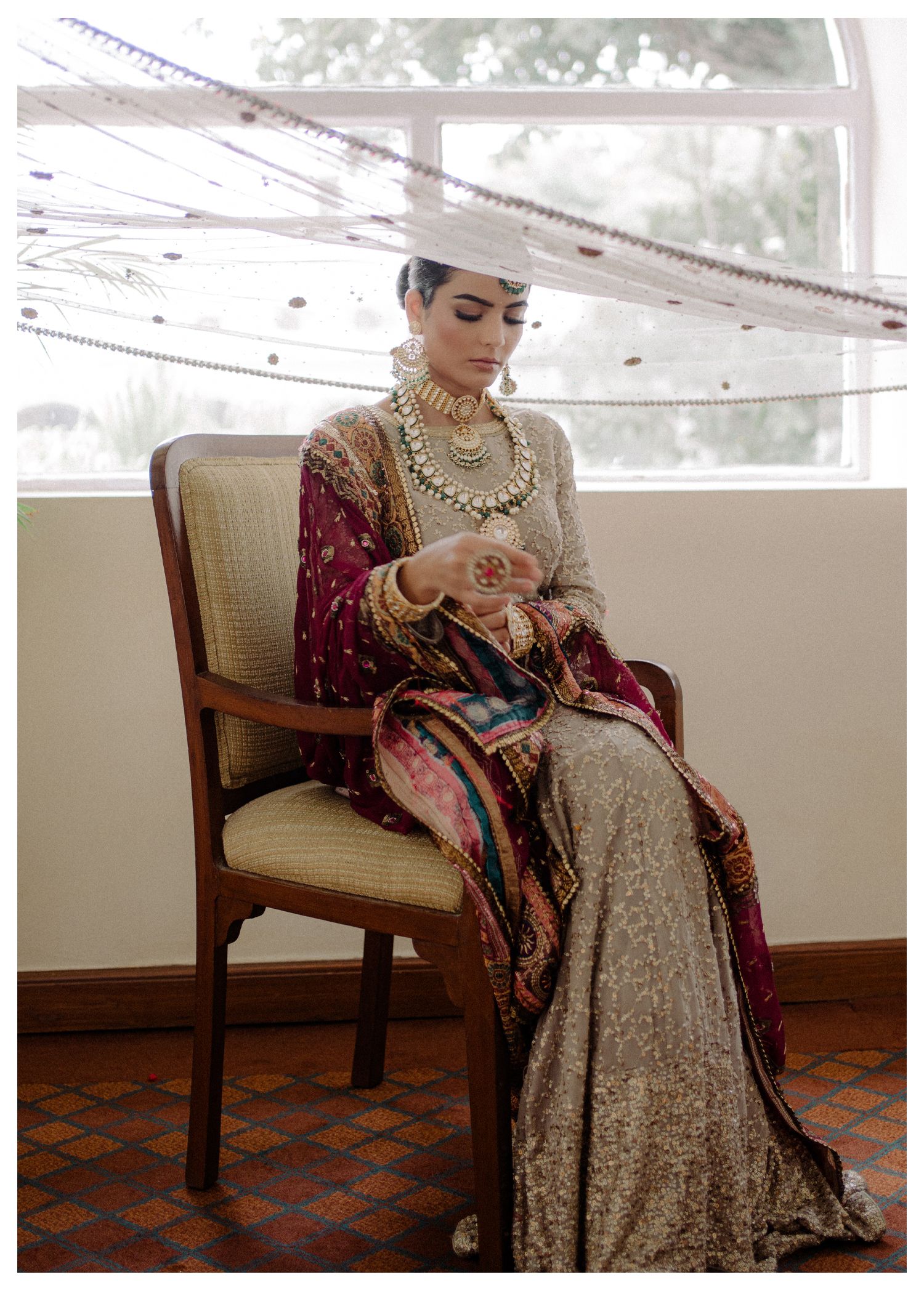 Elegant traditional bridal attire featuring silver embroidery, burgundy velvet shawl and ornate jewelry on a cushioned chair.