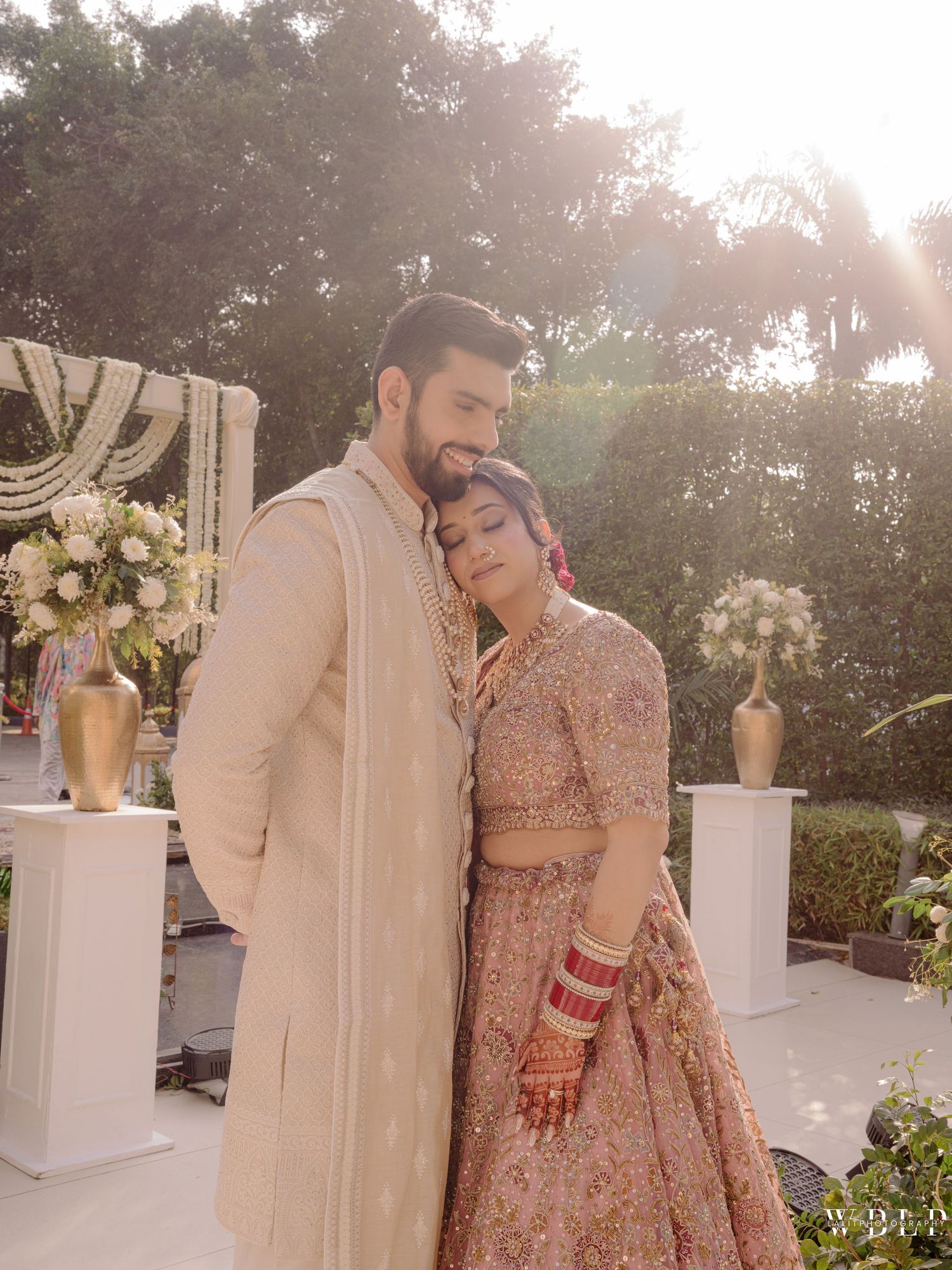 Indian bride and groom in traditional wedding attire pose together outdoors, bride in pink lehenga, groom in cream sherwani.
