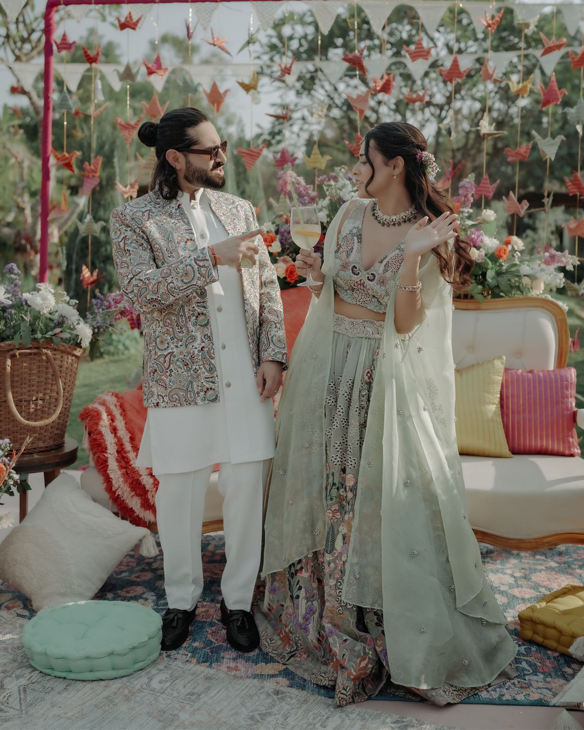 Couple in elegant white and mint Indian wedding attire pose together at an outdoor garden celebration with pink florals.
