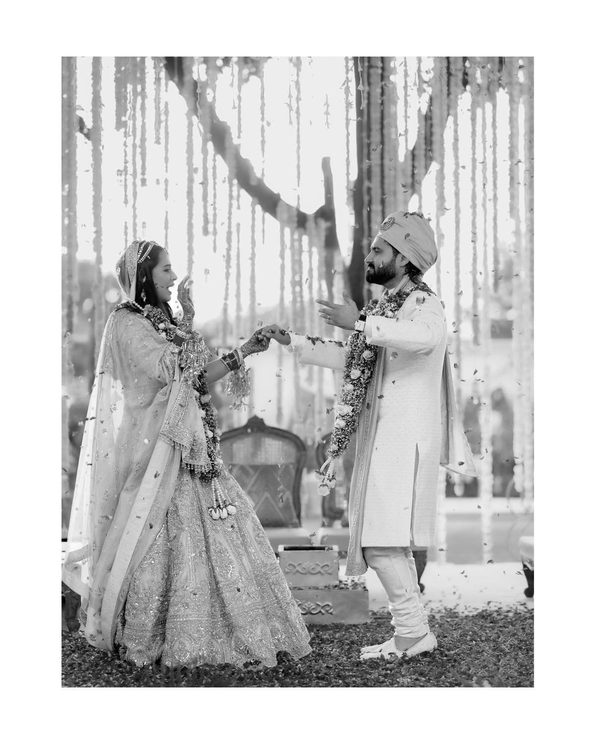 Black and white photo of traditional Indian wedding ceremony with couple exchanging garlands under decorative canopy.