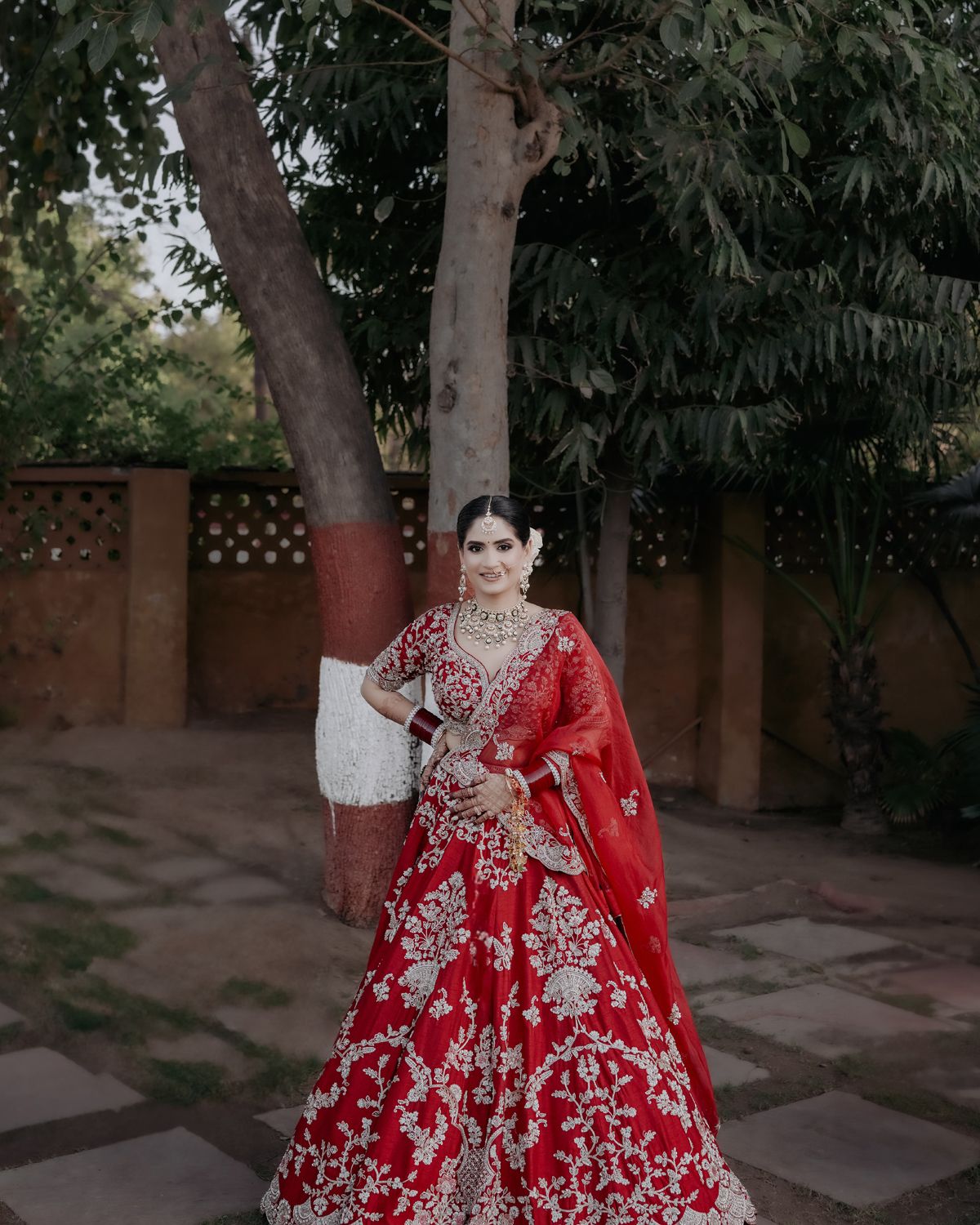 A bride in a stunning red and white embroidered lehenga poses in an outdoor courtyard setting.