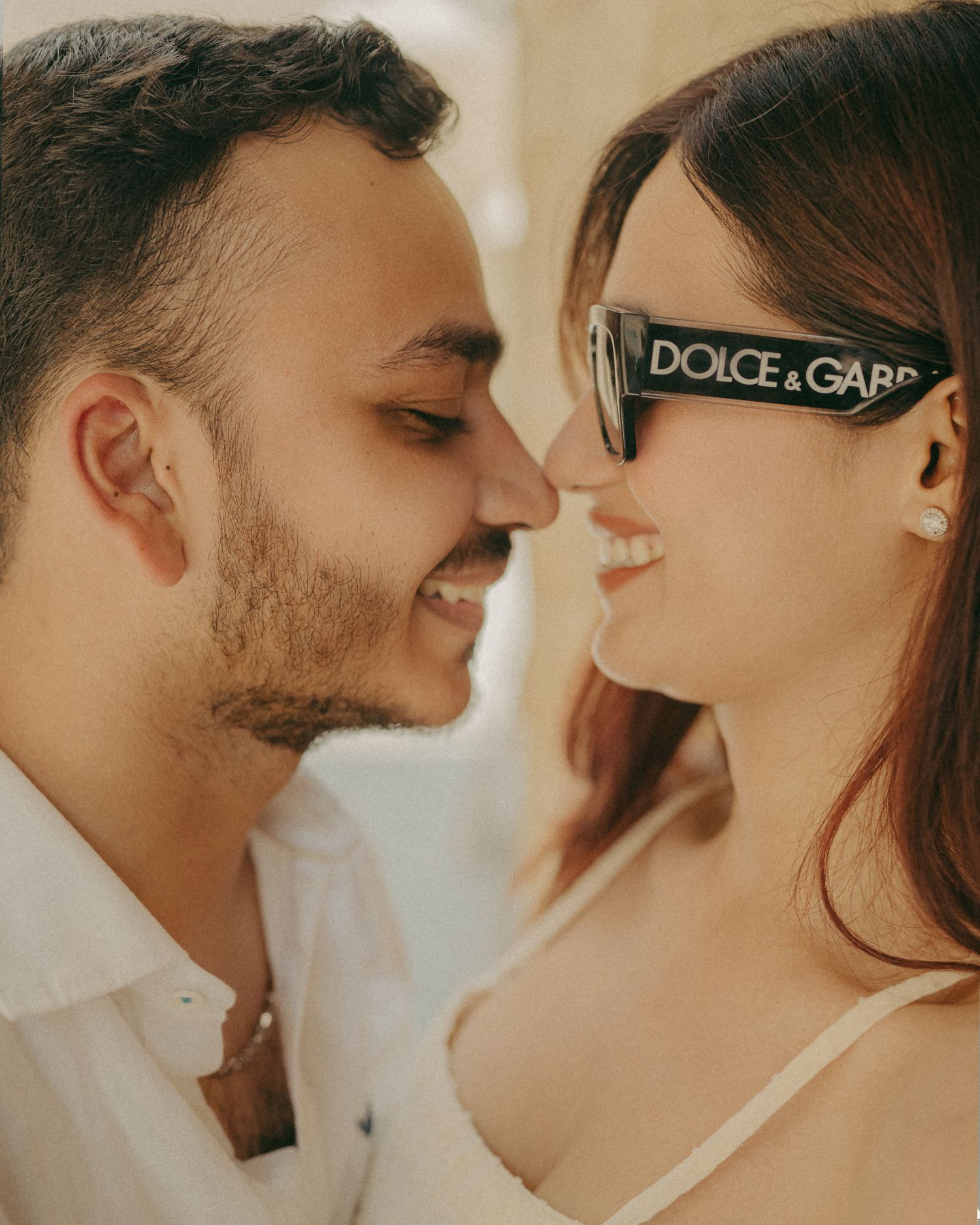 Couple sharing a romantic moment, woman wearing Dolce & Gabbana sunglasses, man in white shirt smiling closely.