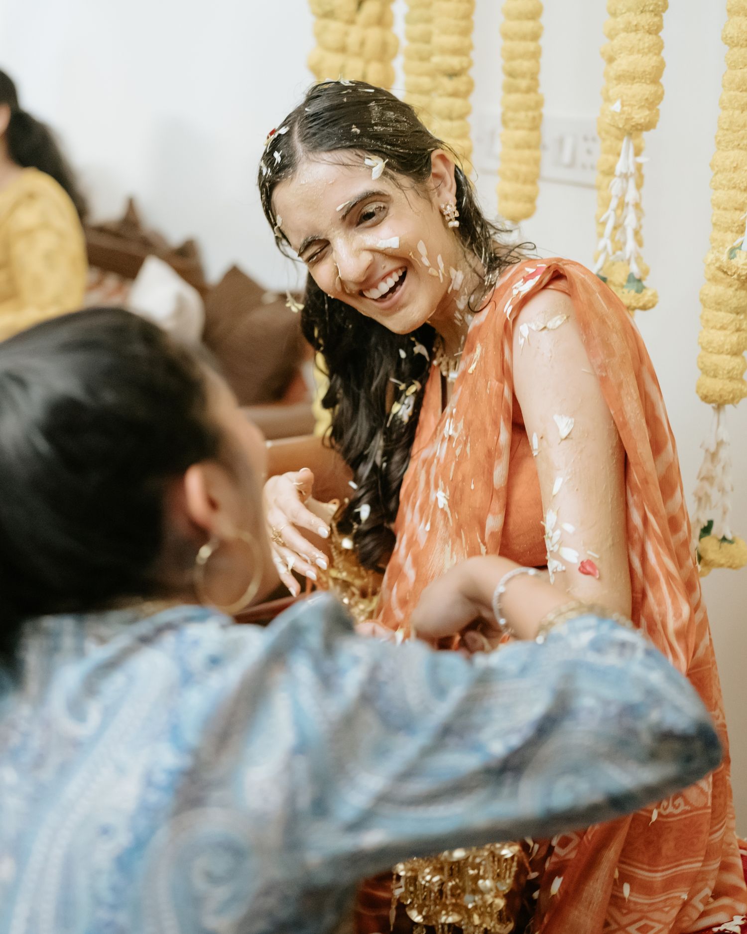 Bride in orange lehenga laughing joyfully during haldi ceremony with flower petals, traditional Indian wedding ritual.