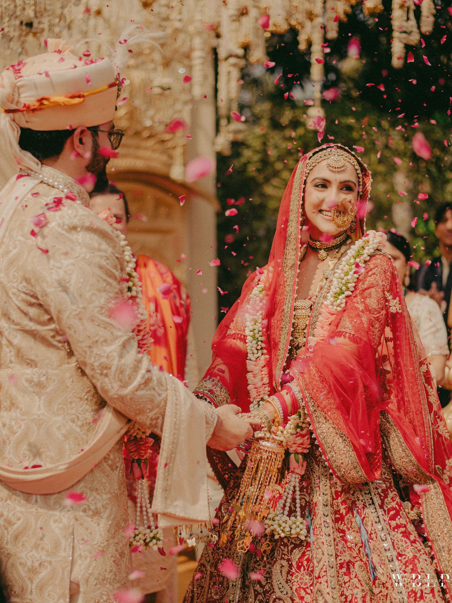 A joyful Indian bride in red lehenga and groom in cream sherwani exchange garlands amid flower petals at wedding.
