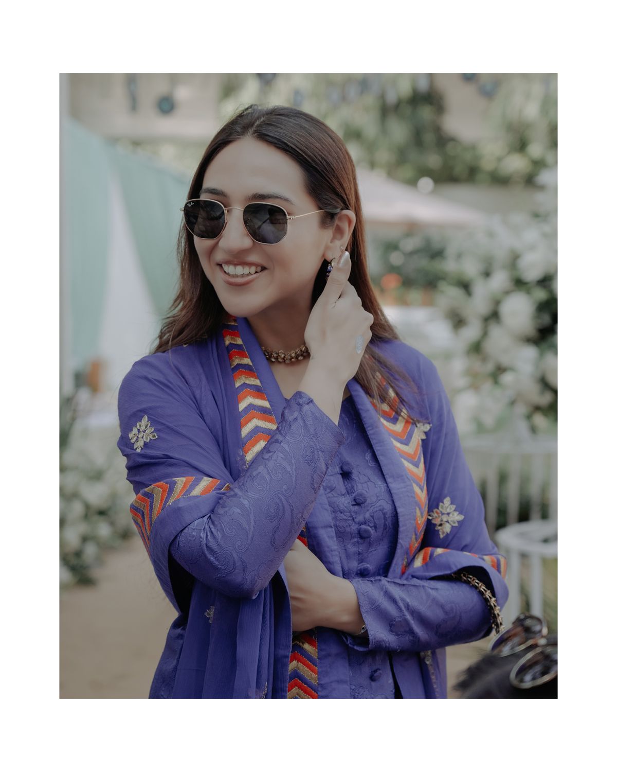 Woman in blue embroidered outfit with sunglasses smiling at an outdoor event.