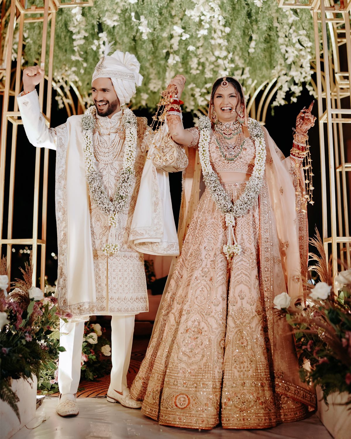 Newlywed couple in matching cream and peach outfits celebrating under floral arch with raised hands.