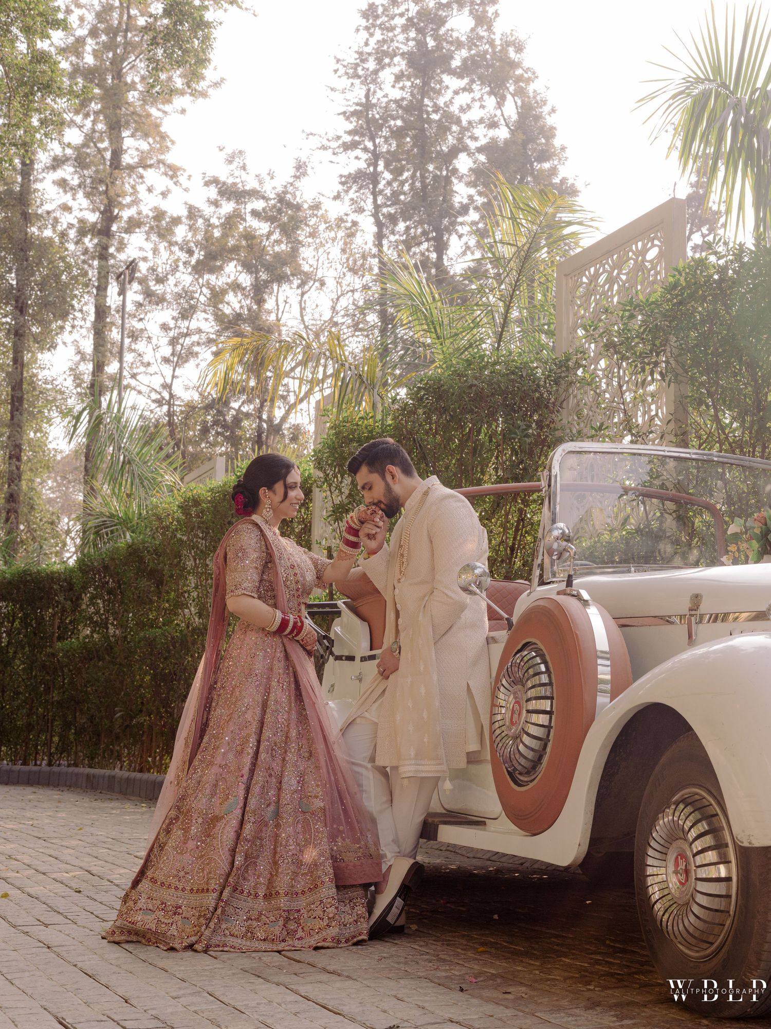 Bride in pink lehenga and groom in cream sherwani pose by vintage white car at outdoor Indian wedding.