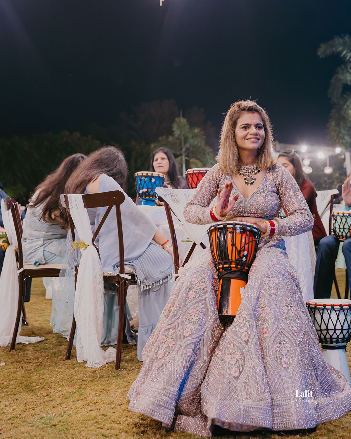 Our Bride in an ornate lace dress plays a drum during an outdoor evening celebration.