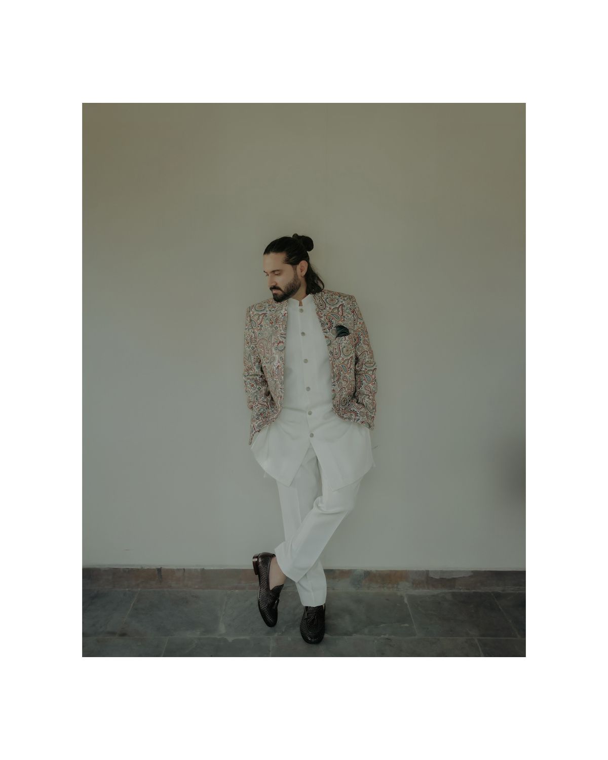 Fashion portrait of a stylish person wearing a patterned blazer and white pants leaning against a neutral wall.