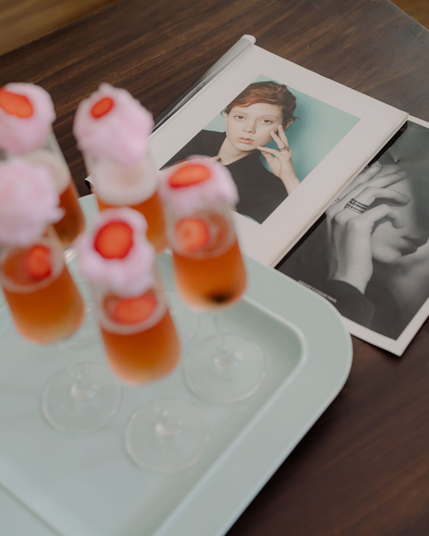 A tray of pink champagne cocktails garnished with strawberries and cotton candy, beside fashion magazine photos on a wooden table.