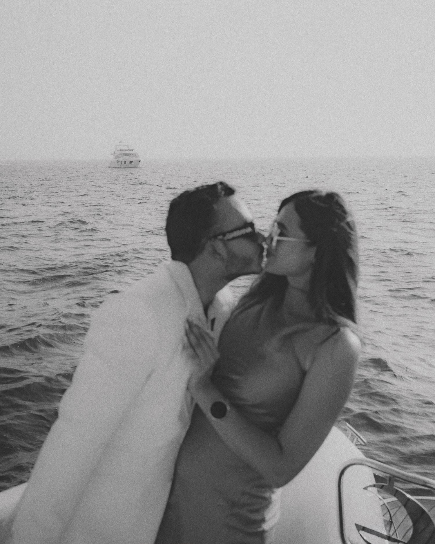 Black and white photo of a couple sharing a romantic kiss on a boat with the ocean and a yacht in the background.