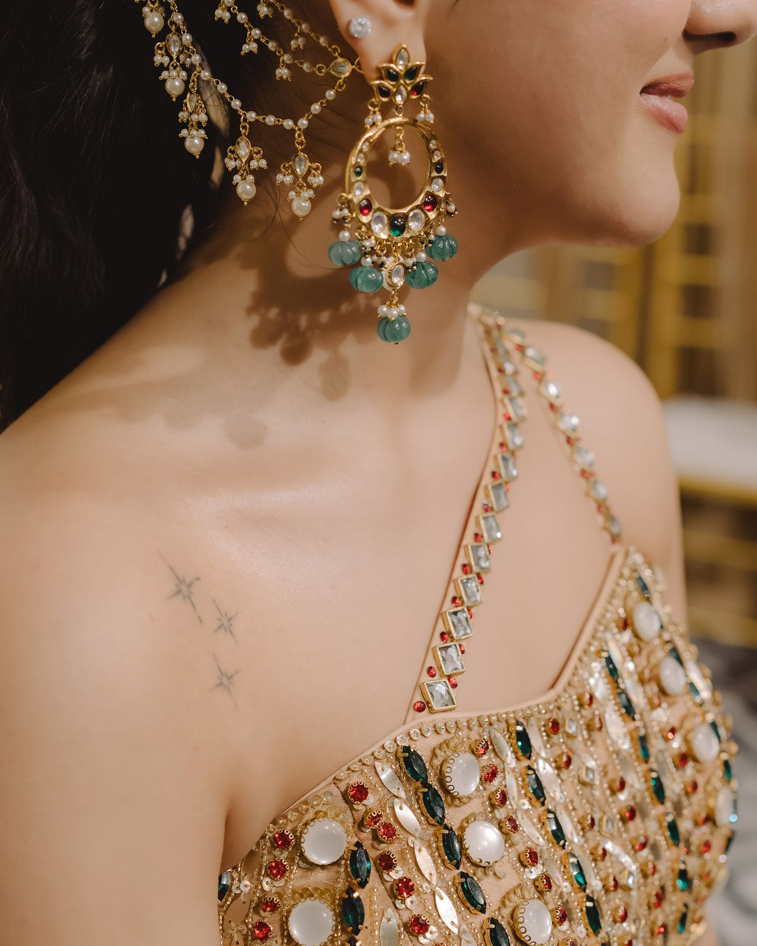 Woman wearing ornate gold chandelier earrings with colorful gemstones, embroidered lehenga, and small star tattoo on collarbone.