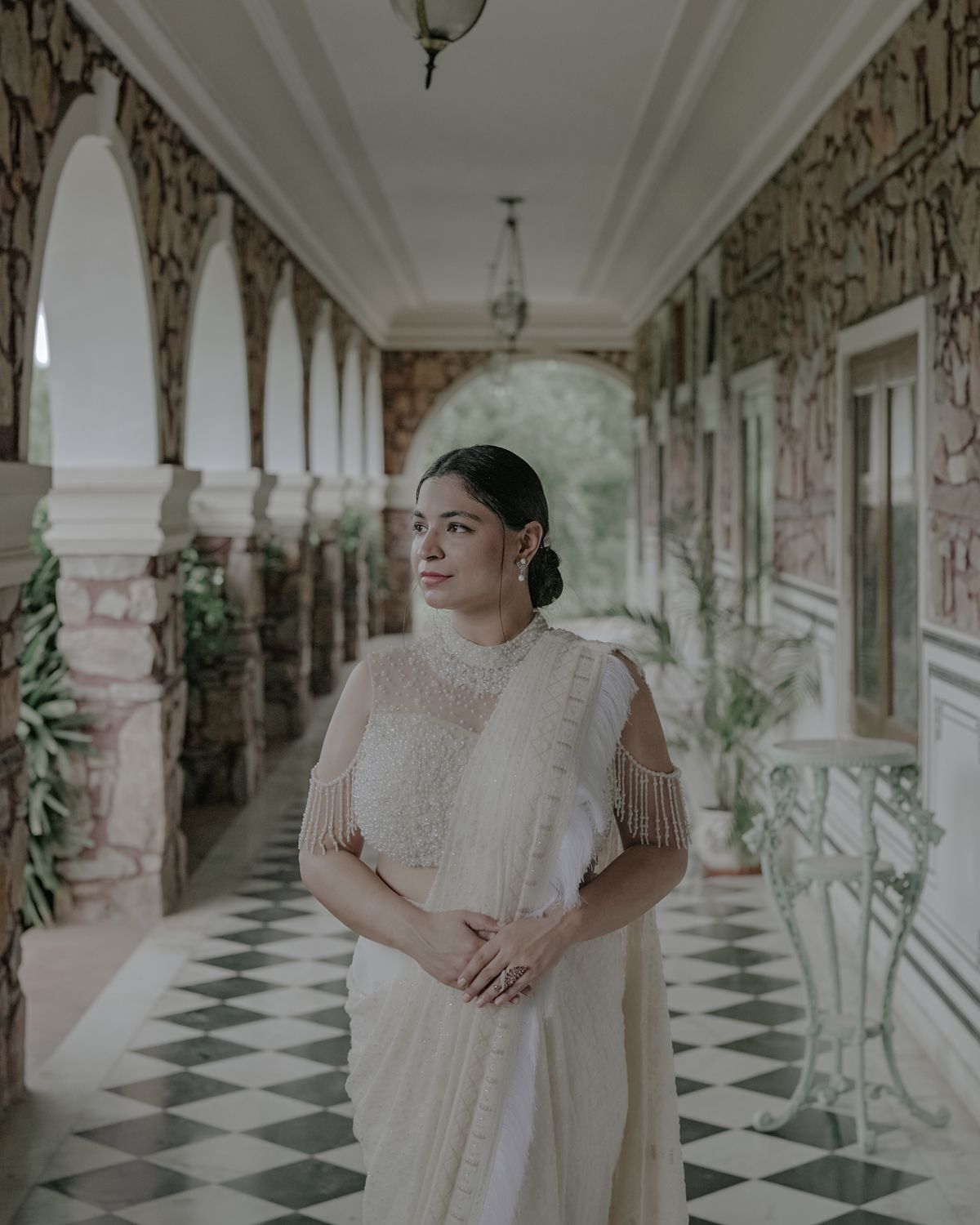 A person in a delicate white saree stands in an ornate corridor with checkerboard flooring and classical architecture.