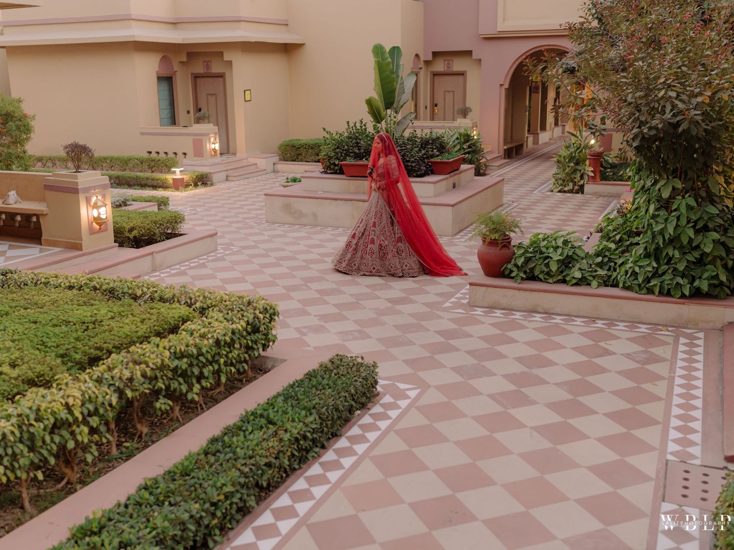 A bride in a red lehenga poses gracefully in an ornate Rajasthani courtyard with manicured gardens and arched architecture.