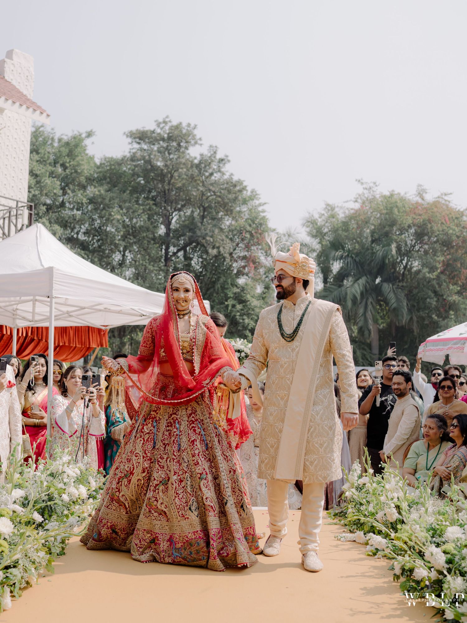 Indian bride in red lehenga and groom in ivory sherwani walk together at outdoor wedding ceremony.