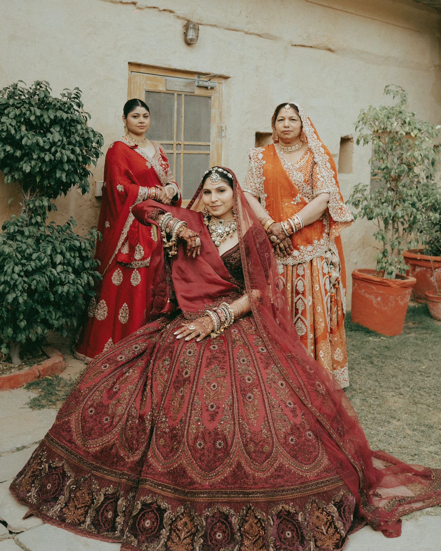 Indian bride in red lehenga with intricate embroidery poses with two women in traditional attire in a courtyard.